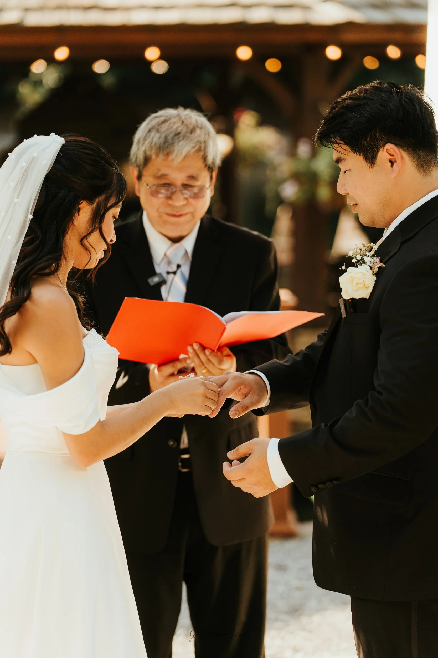 A bride and groom exchange rings during their wedding ceremony, with an officiant reading from a book behind them under outdoor lighting.