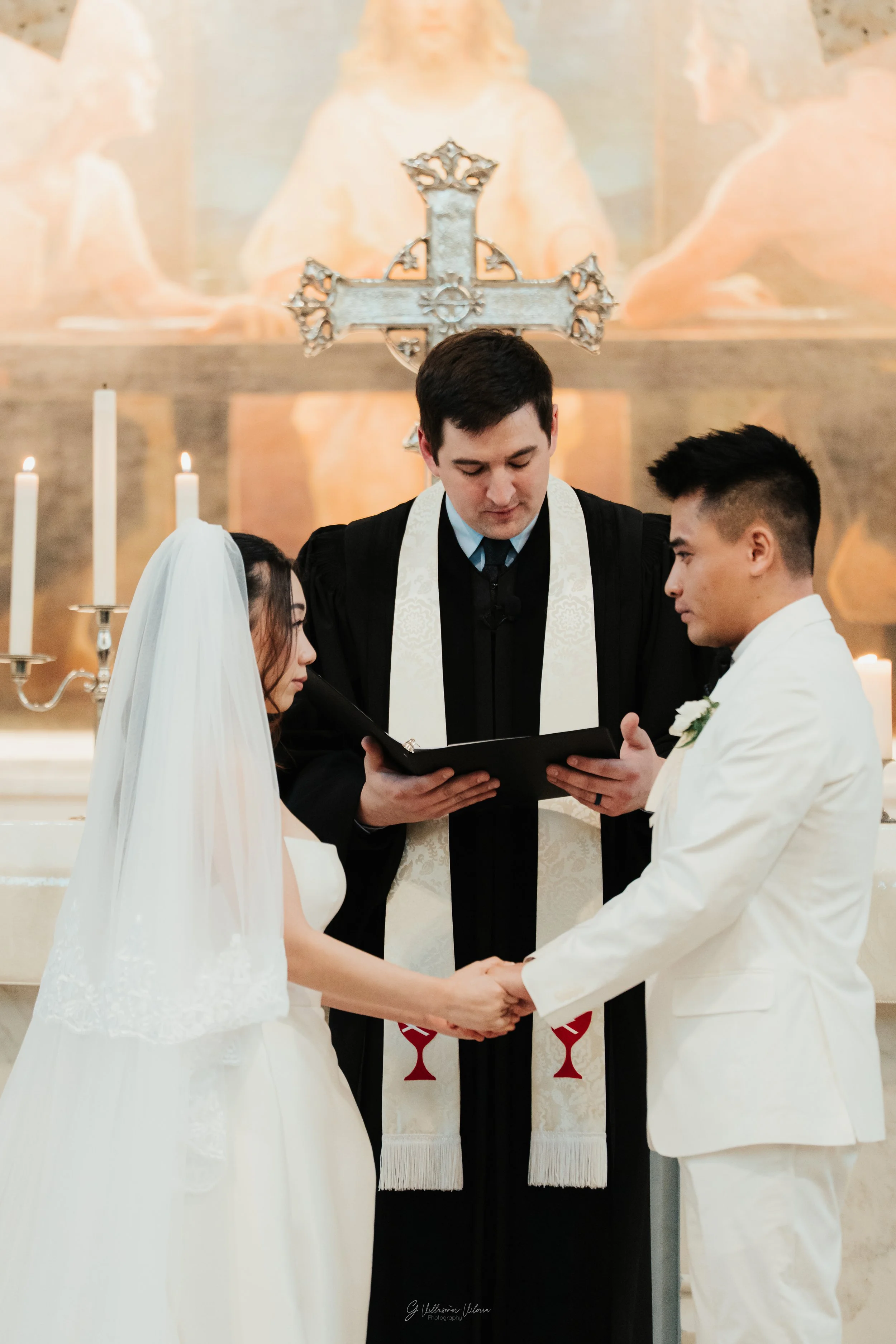 A couple during their wedding ceremony, holding hands and facing each other, with a minister reading from a book. The bride wears a white wedding dress and veil, while the groom wears a white suit. The background shows religious symbols and lit candl