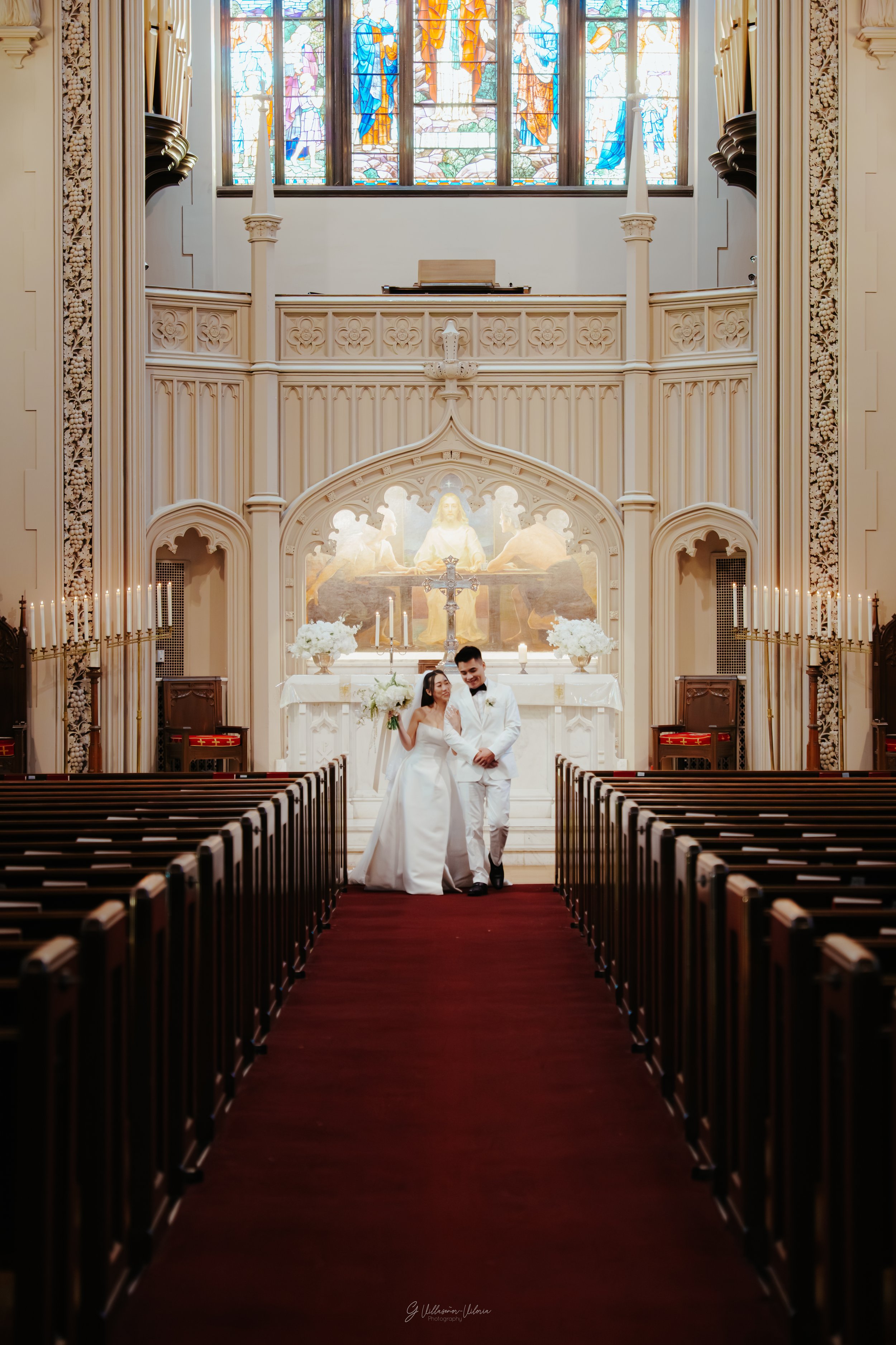 A bride and groom in wedding attire standing inside a church with stained glass windows and ornate architecture, under an altar featuring religious artwork.