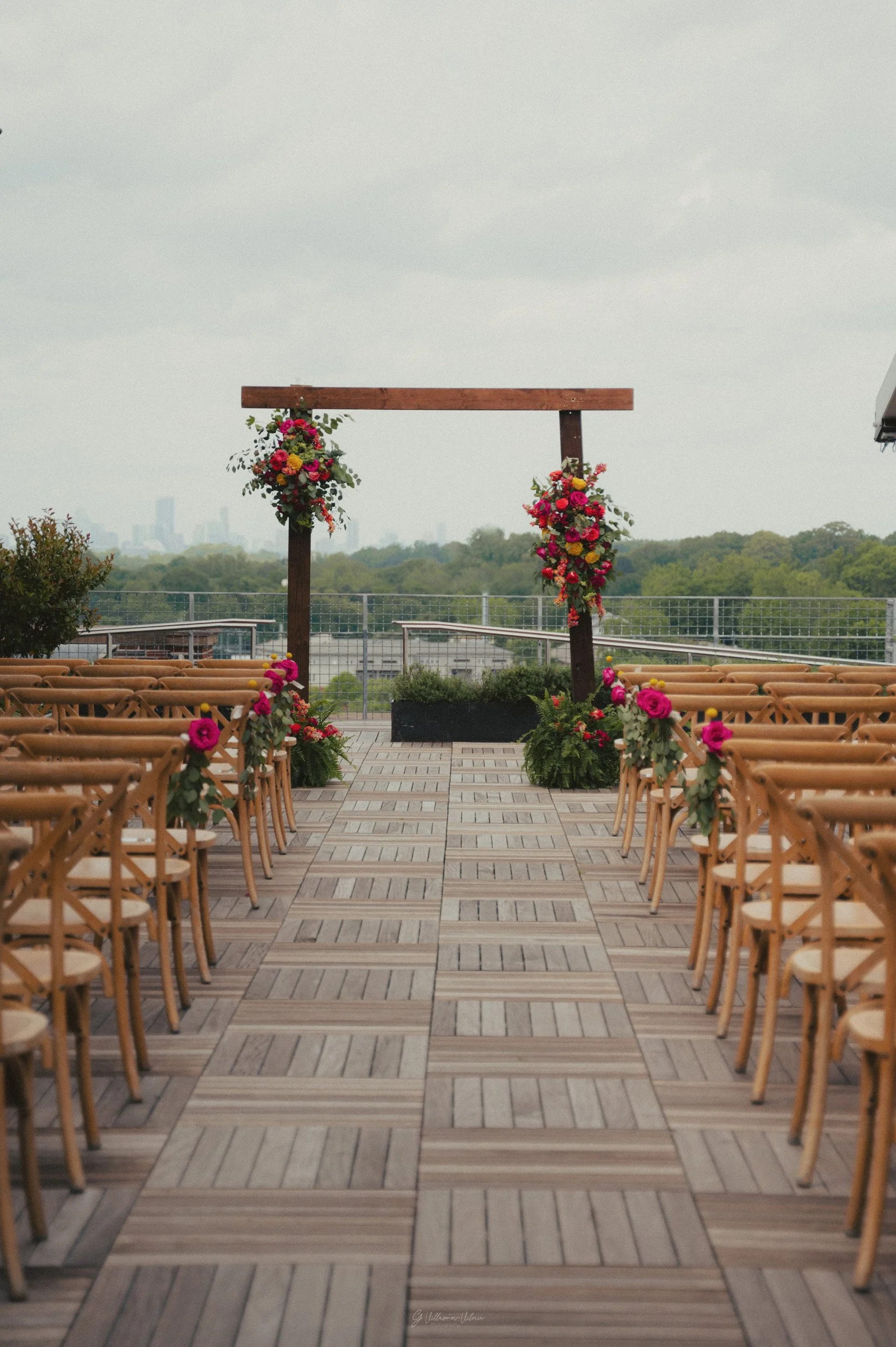 Wedding ceremony setup with wooden chairs on either side of an aisle, decorated with pink and green floral arrangements, leading to a wooden arch adorned with vibrant flowers, on a rooftop with city skyline in the background.