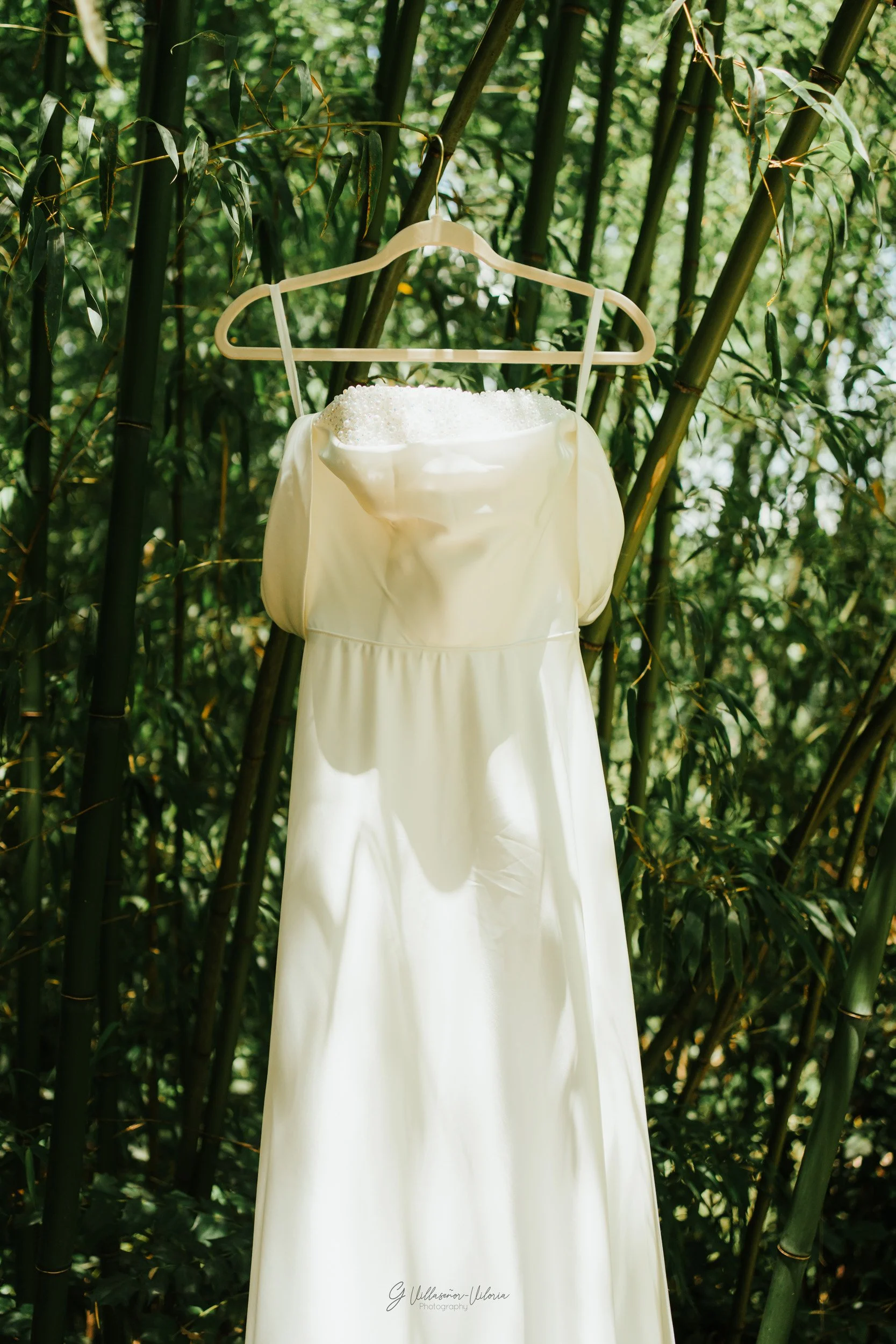A cream-colored wedding dress hanging on a white hanger, displayed on bamboo stalks in a lush green outdoor setting.