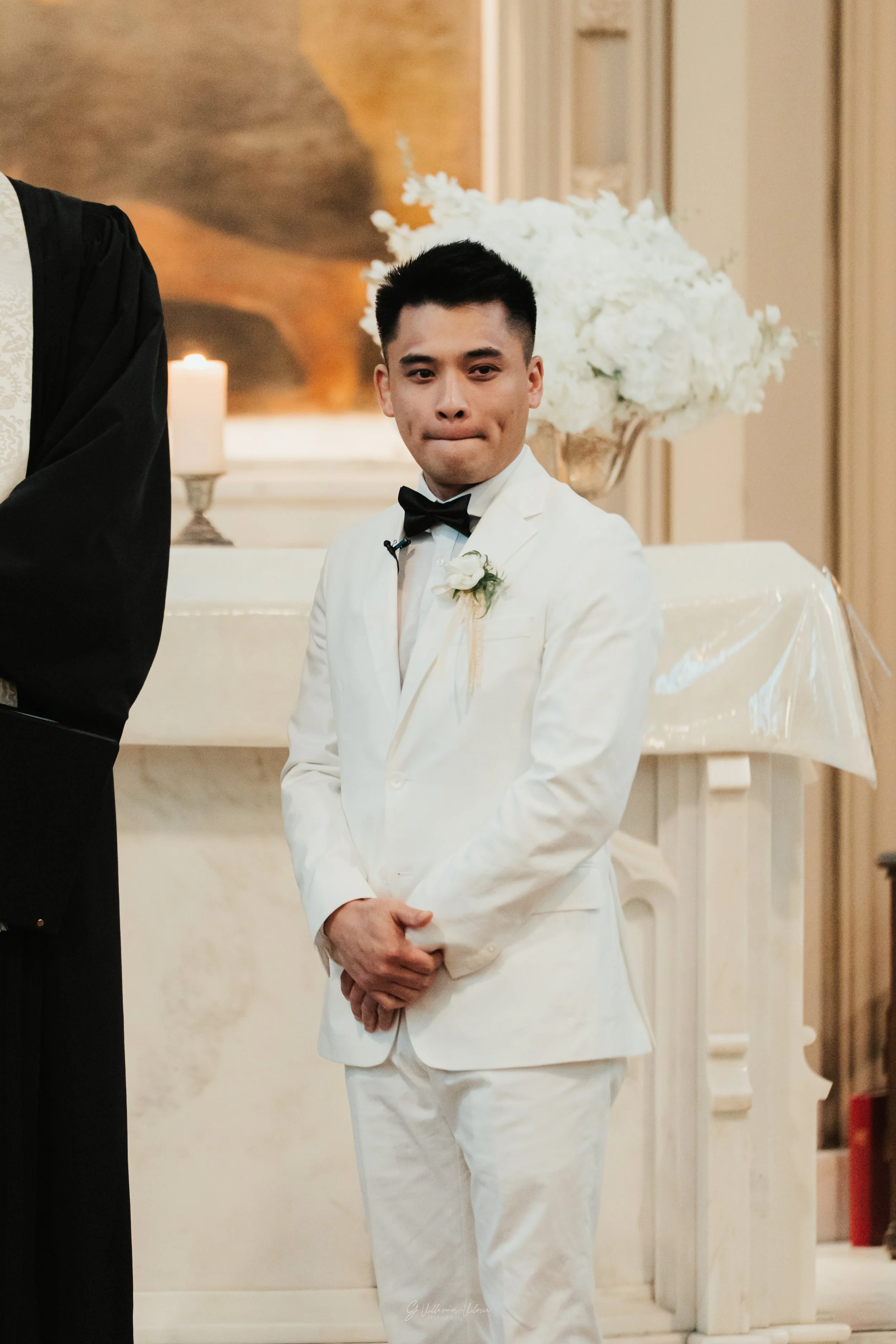 A man dressed in a white tuxedo with a black bow tie, standing with his hands clasped in front of him, in a decorated interior setting with a large white floral arrangement and candles.