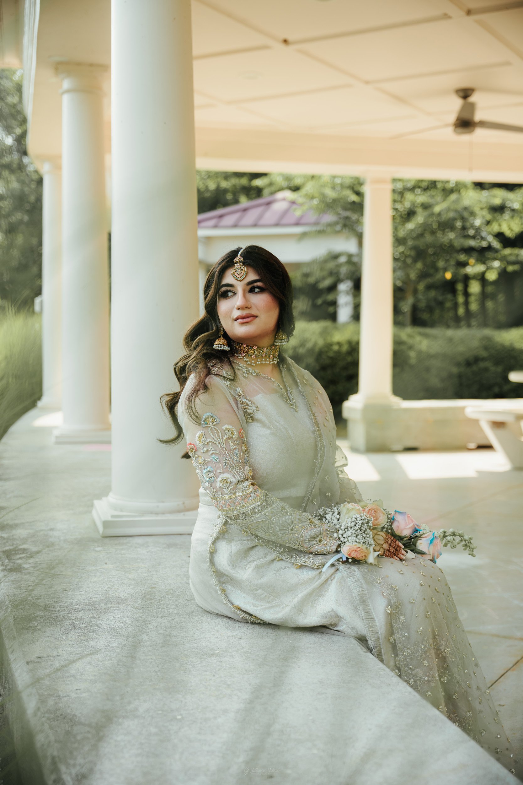 A woman dressed in traditional Indian wedding attire sits on a stone bench outdoors, holding a bouquet of flowers, with a white pavilion and greenery in the background.
