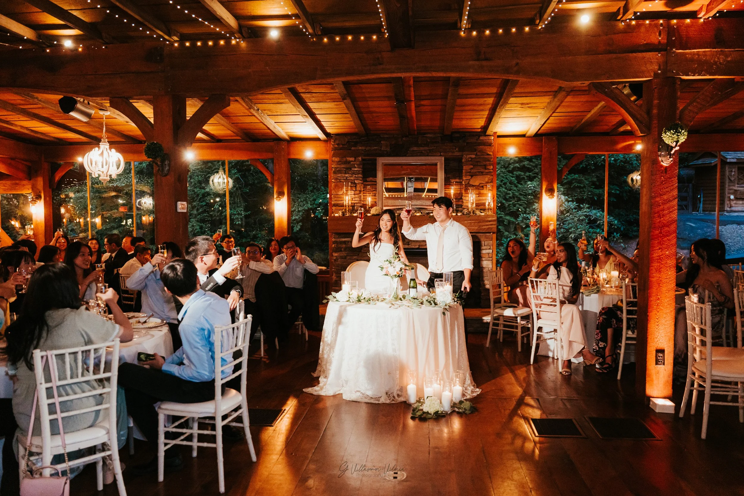 A wedding celebration in a warmly lit, rustic wooden venue. The bride and groom stand behind a table, raising glasses in a toast, surrounded by seated guests who are also raising their glasses. The scene is decorated with candles and flowers, with la