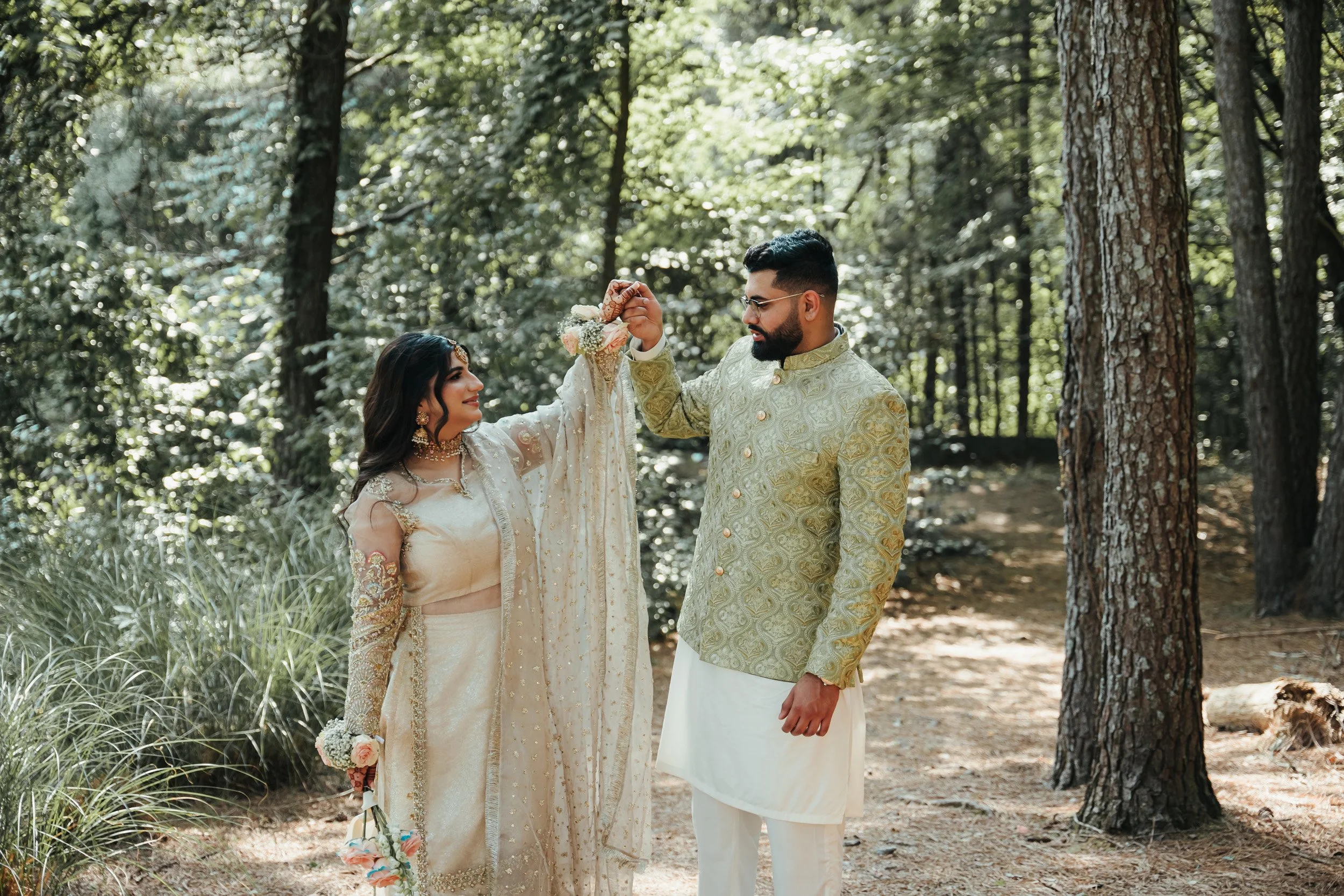 A couple dressed in traditional South Asian wedding attire stand in a forest, with the woman holding a small bouquet and the man holding a floral accessory.
