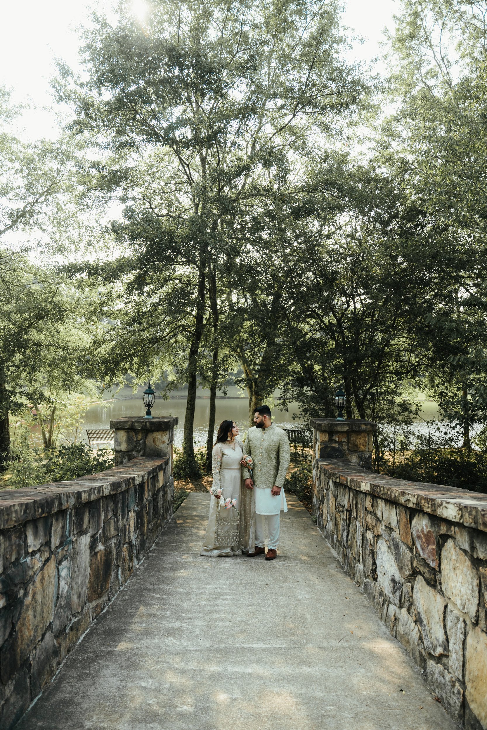 A couple dressed in traditional wedding attire standing on a stone bridge by a lake, surrounded by trees.