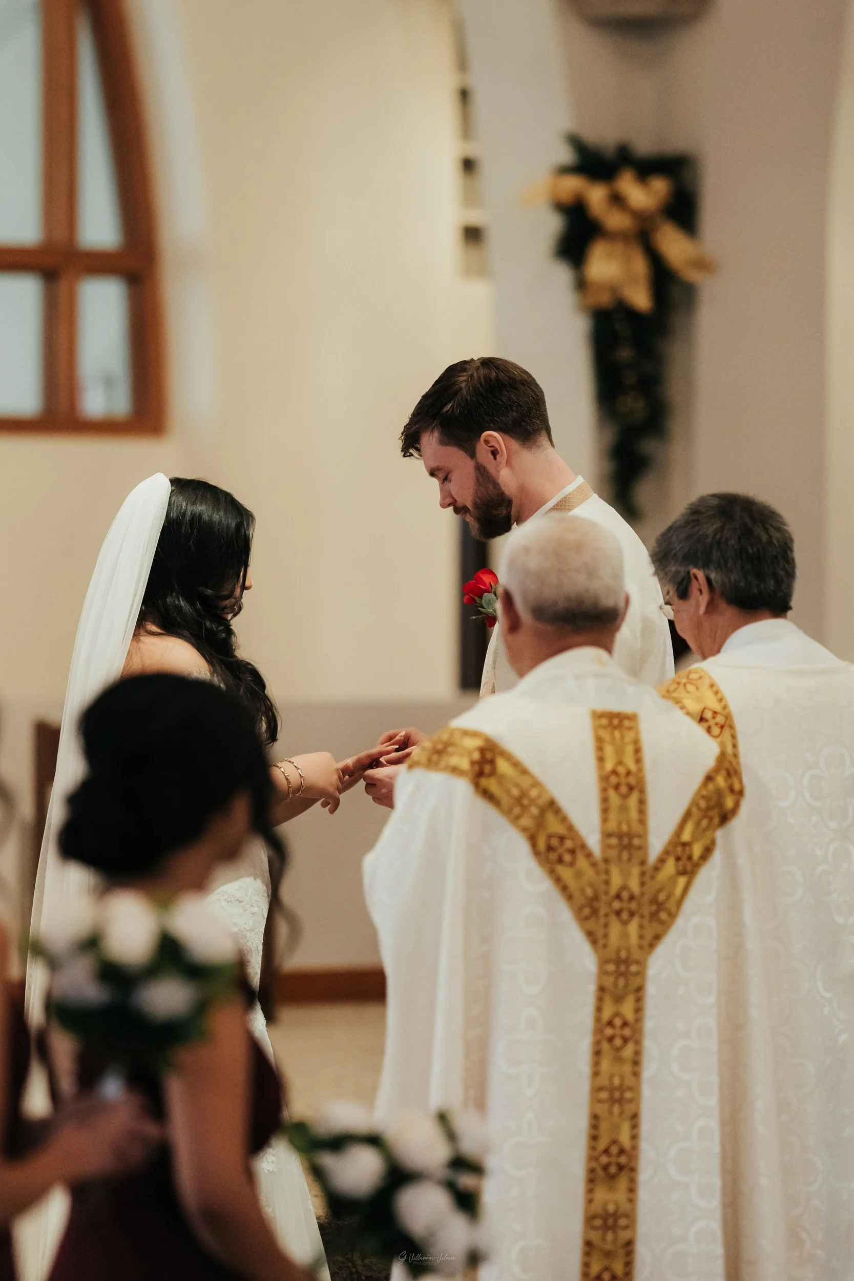 A wedding ceremony in a church with a priest, a bride with a white veil, a groom, and floral arrangements.