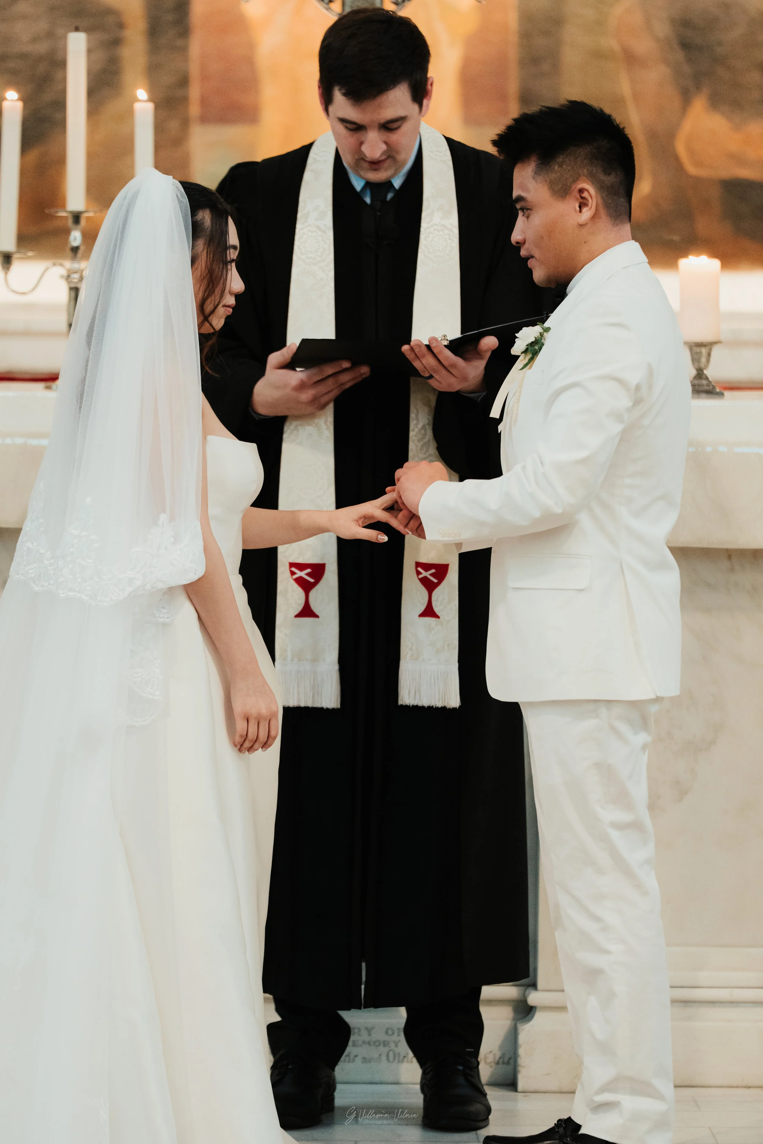 A couple getting married at an indoor church ceremony, with a priest officiating, candles in the background, and the couple holding hands during the exchange of vows.