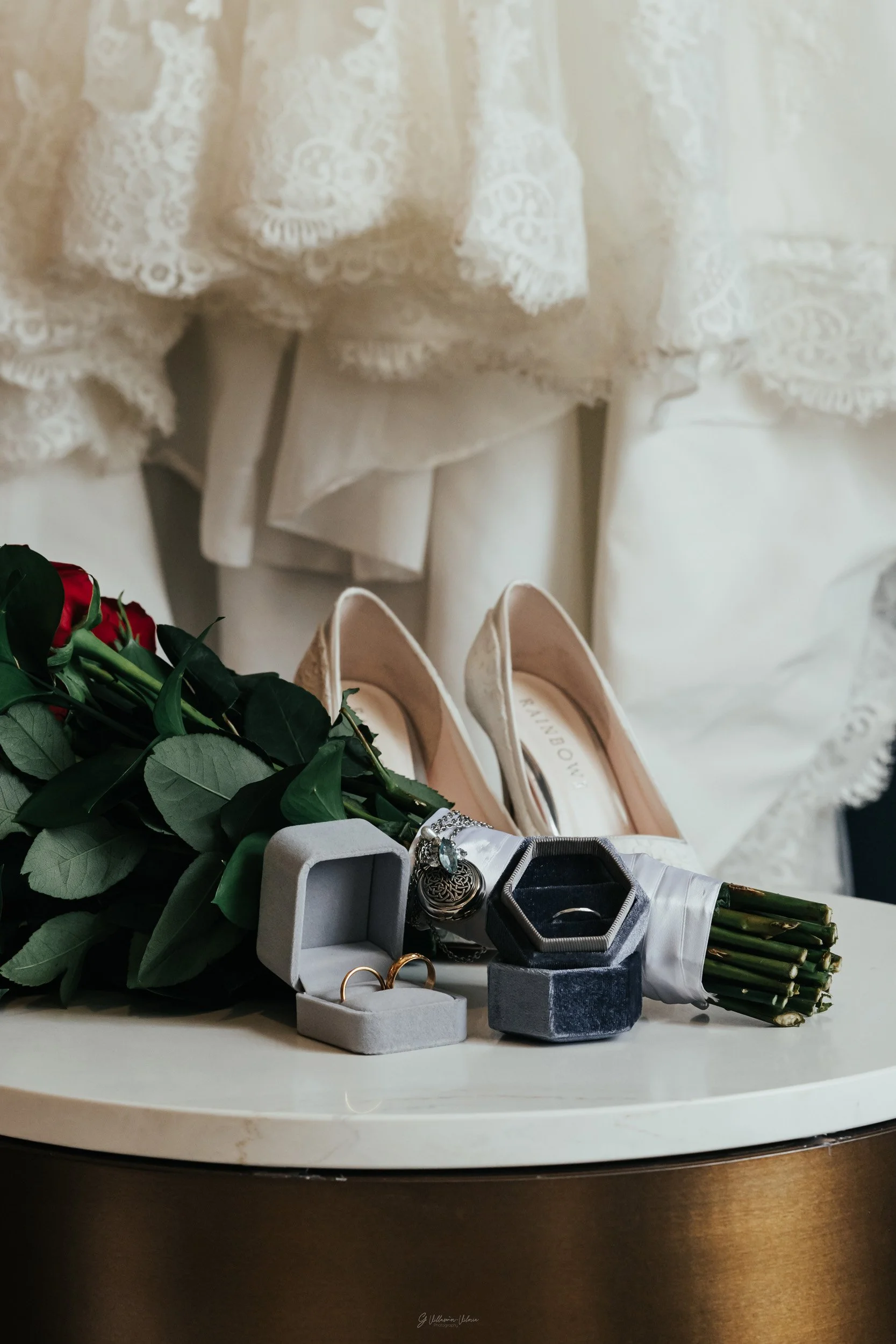 A wedding dress with lace details, a bouquet of red roses, a pair of beige high-heeled shoes, and jewelry including rings and bracelets on a white table.