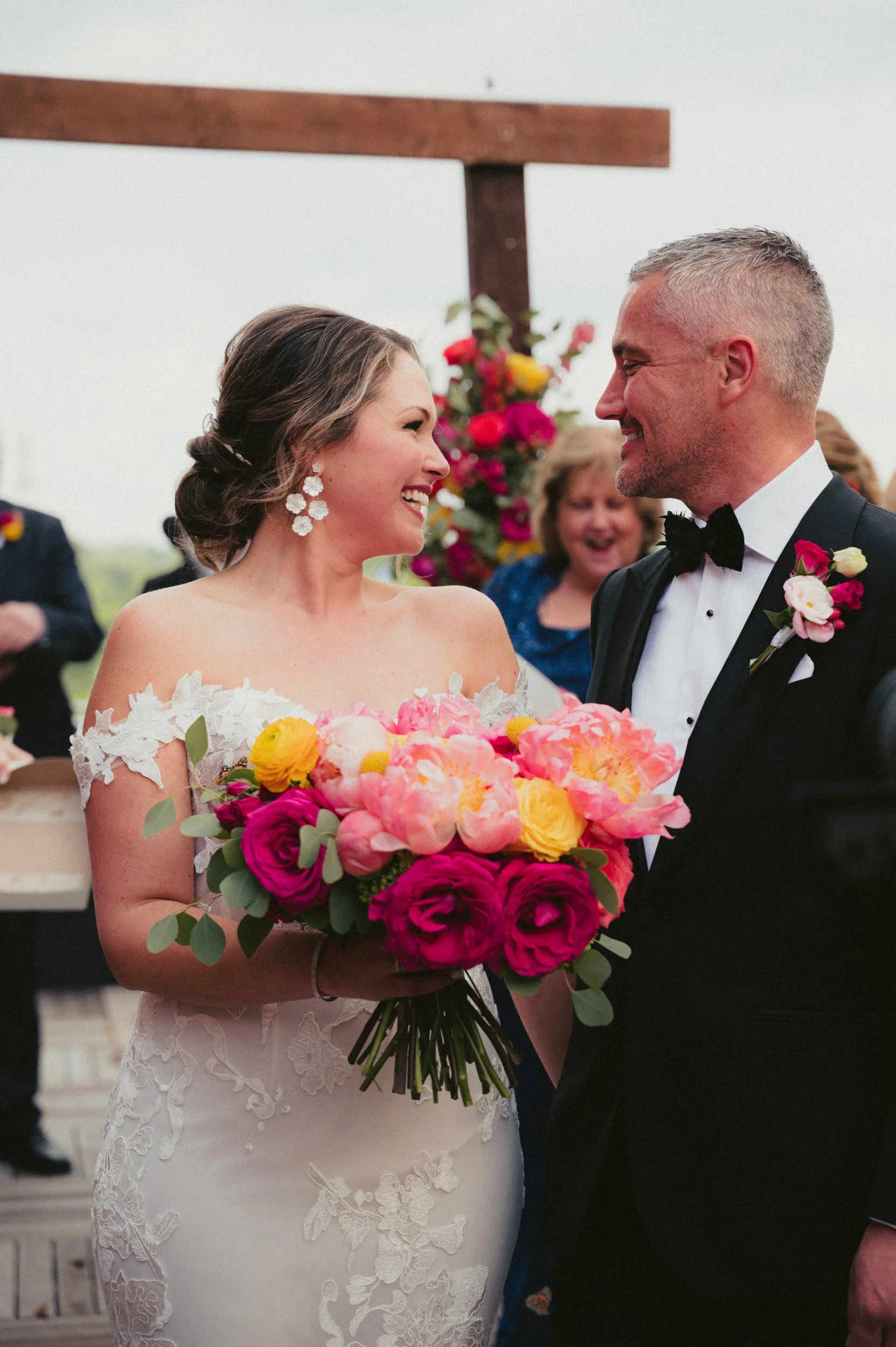 Bride and groom smiling at each other during a wedding ceremony, with the bride holding a colorful bouquet of pink, yellow, and white flowers, and others in the background.