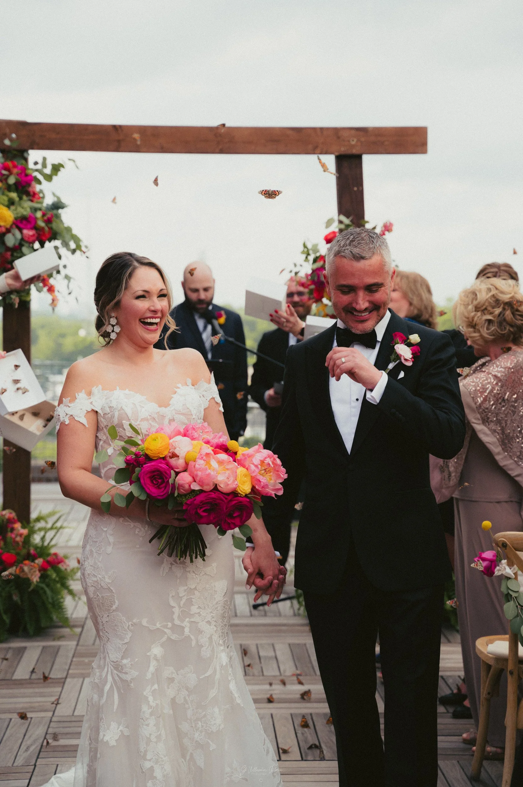 A smiling bride holding a colorful bouquet walks hand-in-hand with a smiling groom in a black tuxedo at their wedding ceremony, with guests and a floral arch in the background.