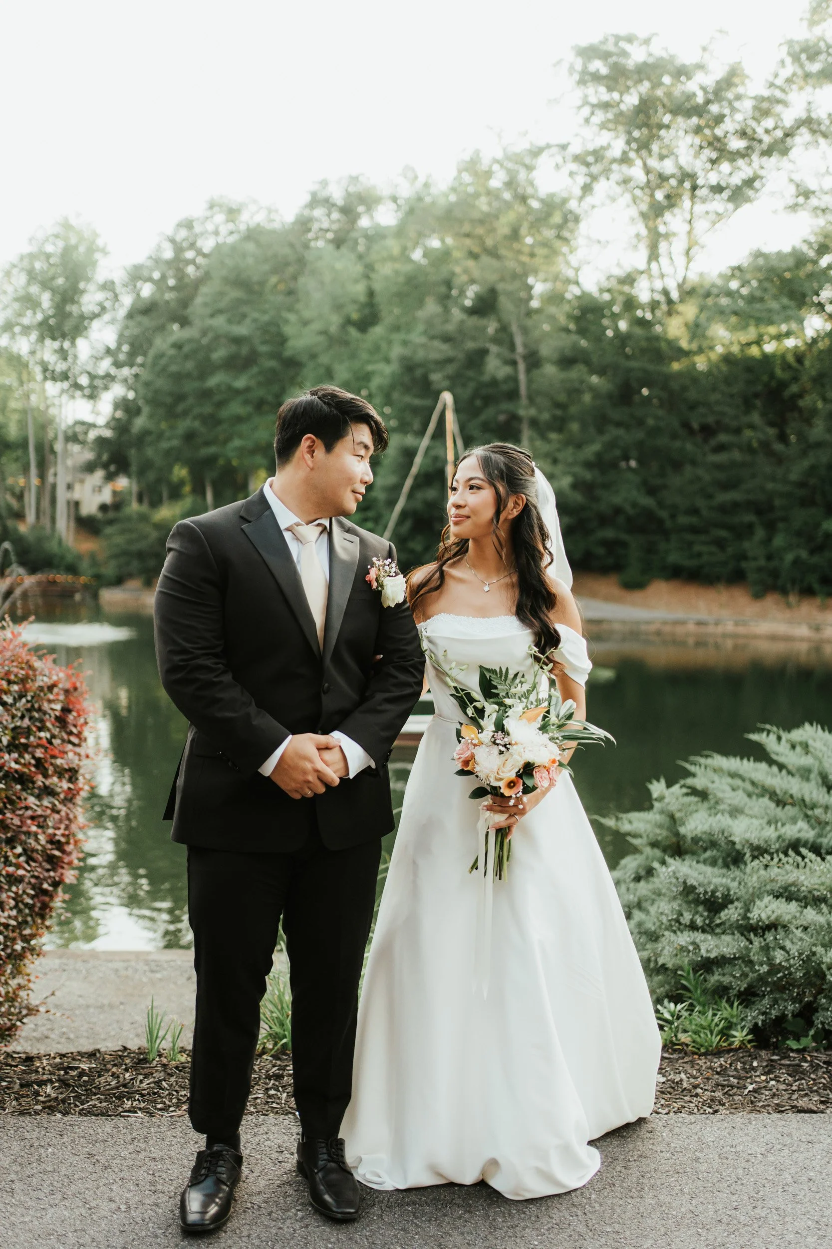 A bride and groom standing together outdoors near a body of water with trees in the background, dressed in wedding attire. The bride is holding a bouquet of flowers and looking at the groom, who is in a black suit and tie.