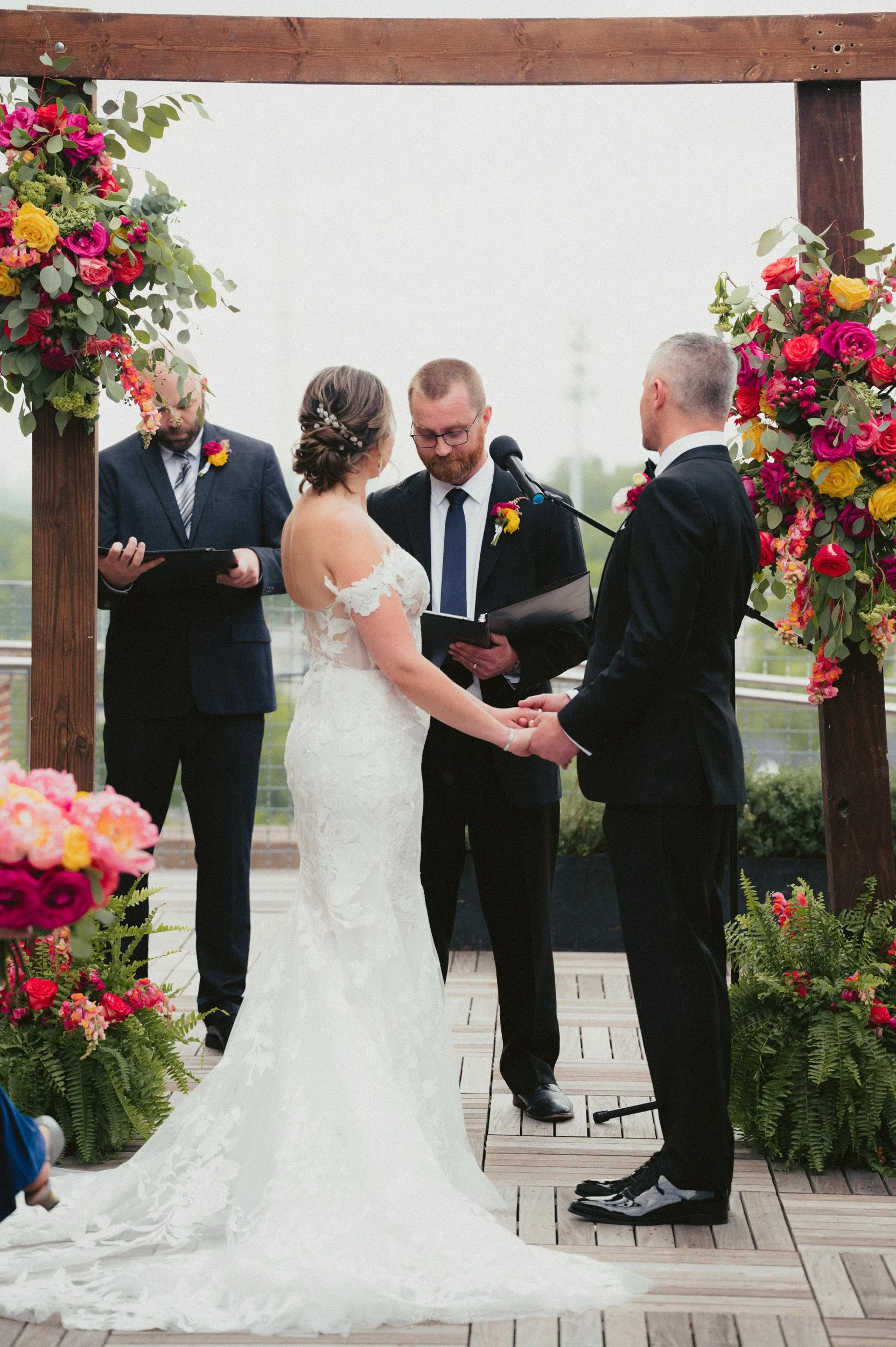 A bride and groom holding hands during a wedding ceremony outdoors, with an officiant and two groomsmen dressed in suits, under a wooden arch decorated with colorful flowers.