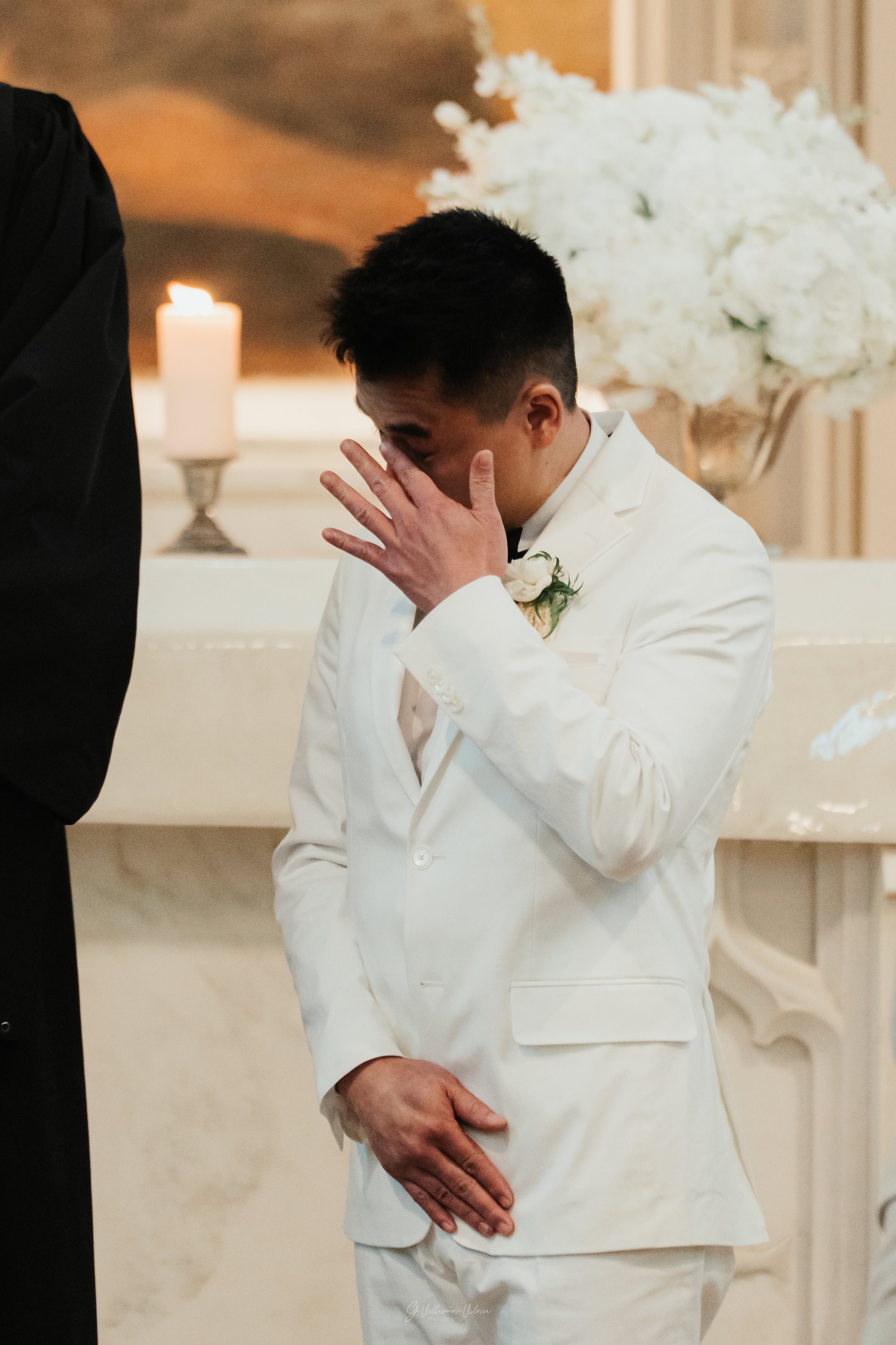 A man dressed in a white suit appears emotional, covering his face with one hand during a wedding ceremony, with white flowers and candles in the background.