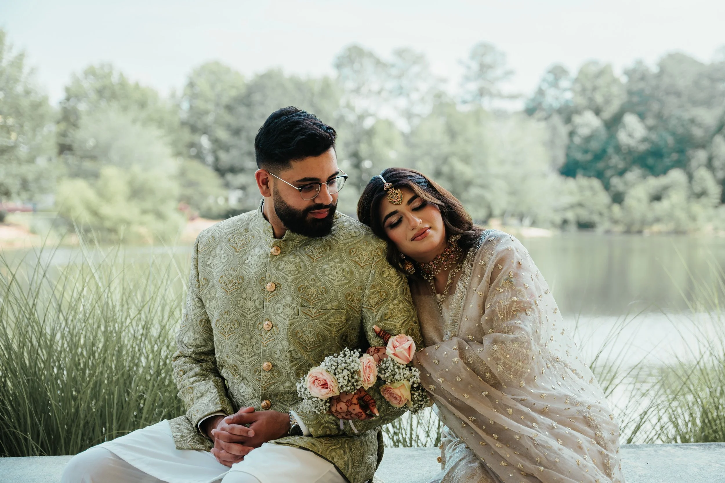 Indian couple dressed in traditional attire sitting by a lake with greenery, holding a bouquet of pink roses, enjoying a peaceful moment.