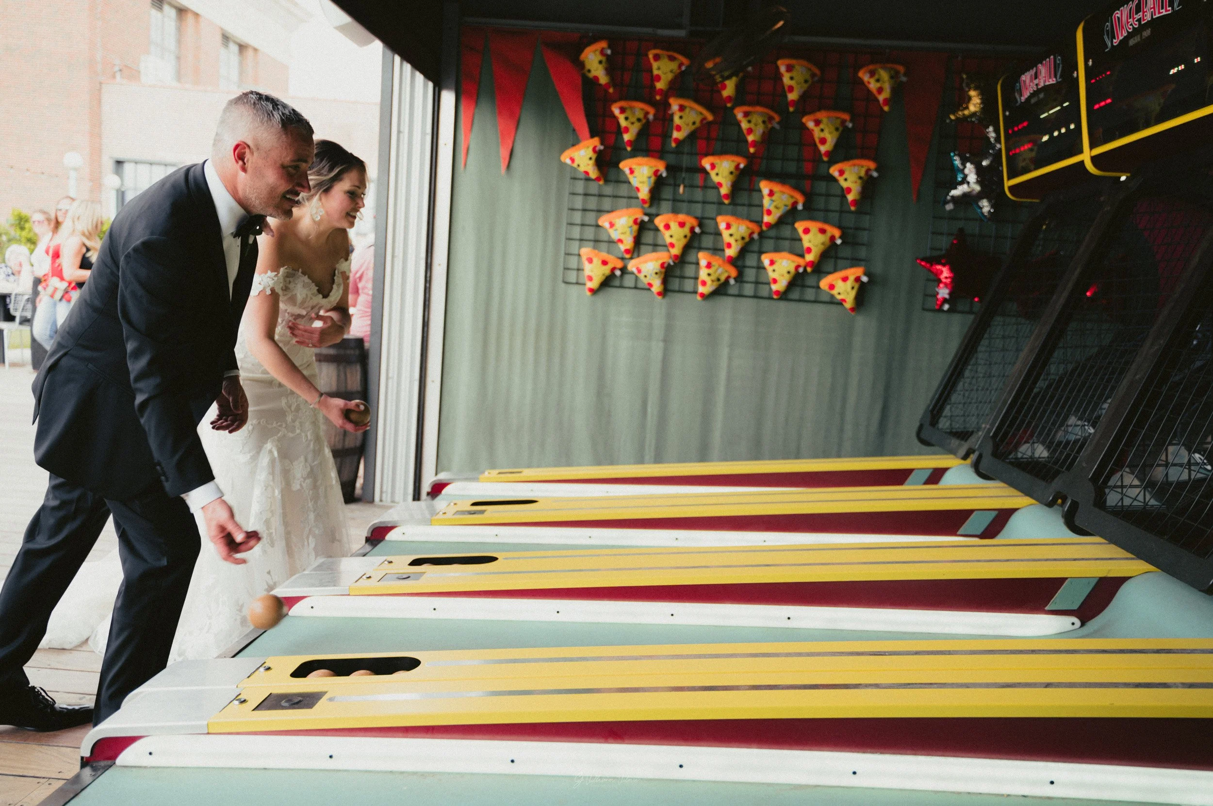 A bride and groom playing skee-ball at an arcade or carnival, with the bride in a white wedding dress and the groom in a black tuxedo, both reaching for balls on the game.