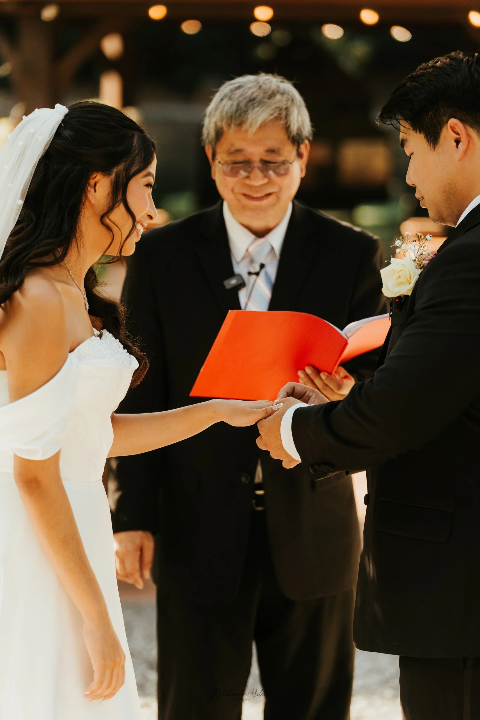A bride and groom holding hands during their wedding ceremony, with a officiant reading from a red book in a warmly lit outdoor setting.