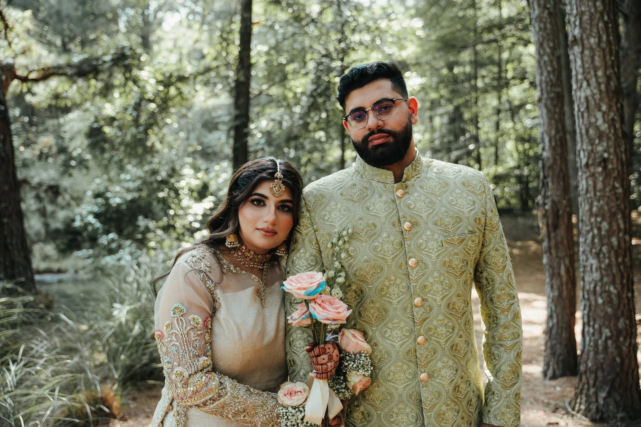 A South Asian couple in traditional wedding attire standing in a wooded outdoor setting. The woman wears a heavily embroidered beige and gold dress with jewelry, including a headpiece and choker, and holds a bouquet of flowers. The man wears a light 