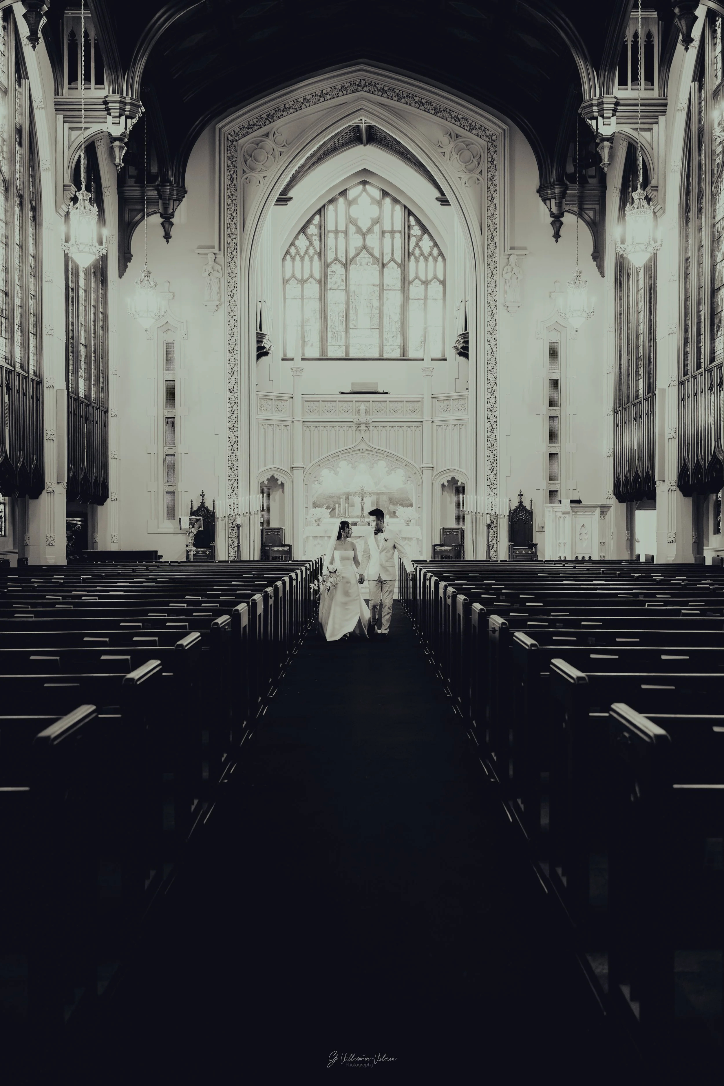 A couple walking down the aisle of a church, holding hands, on their wedding day with high vaulted ceiling and stained glass windows.