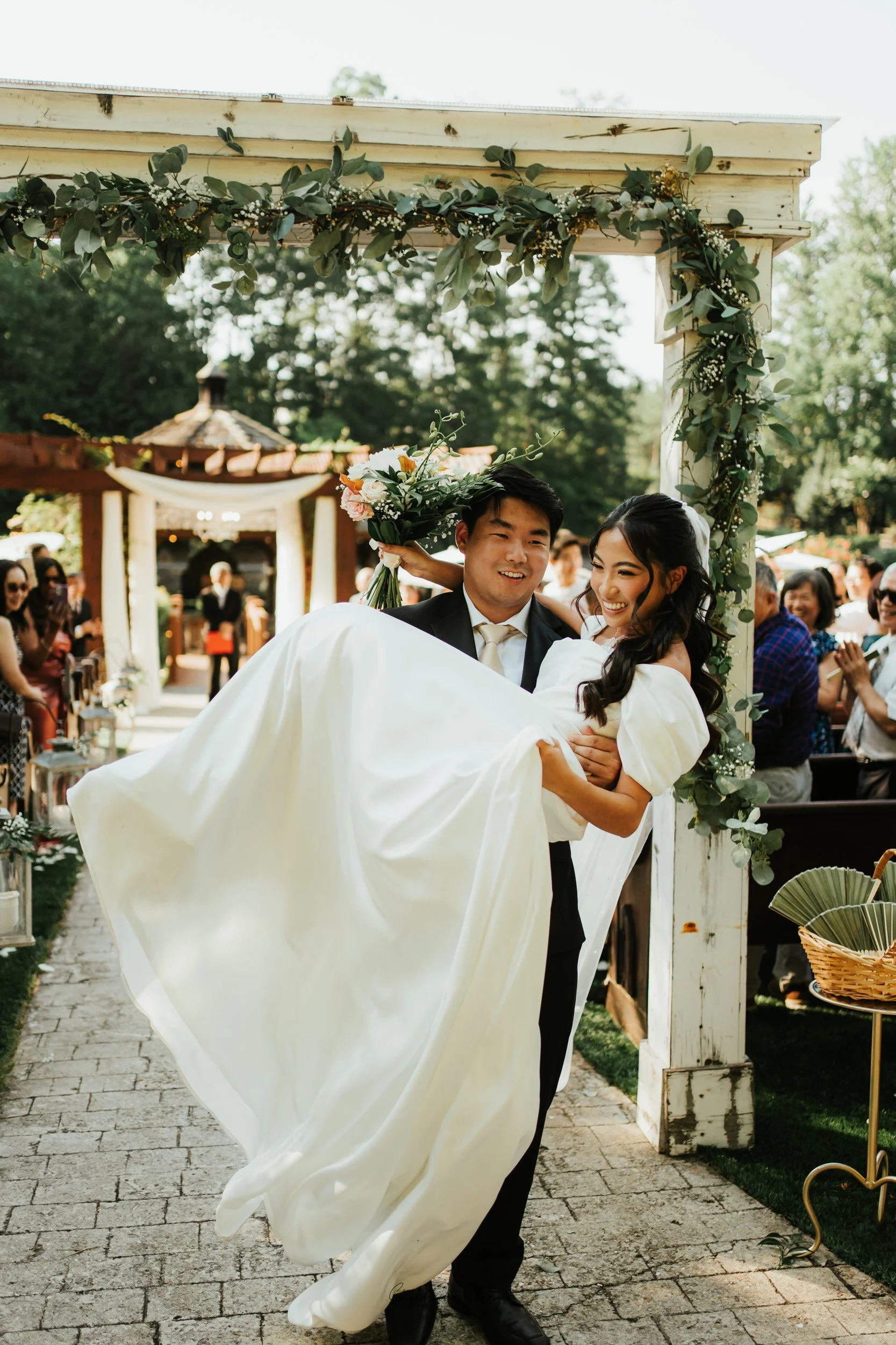 Couple at wedding, groom carrying bride, wedding arch decorated with greenery, guests clapping and smiling in outdoor garden setting.