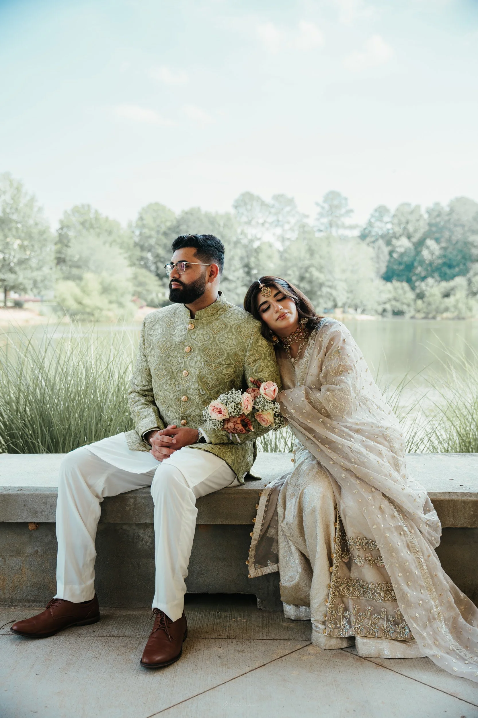 A couple dressed in traditional Indian wedding attire sitting on a bench by a lake with greenery in the background. The woman is resting her head on the man's shoulder and holding a bouquet of pink and white flowers.