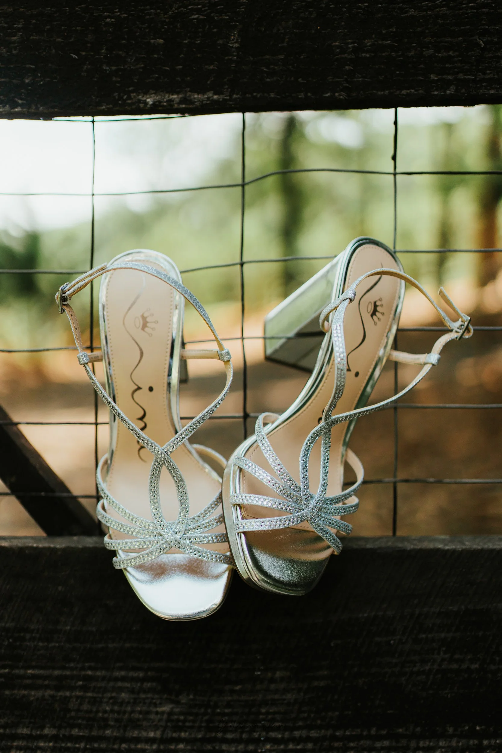 Silver high-heeled sandals with rhinestone embellishments hanging on a black wooden railing with a wire mesh, with a blurred outdoor background of trees and sky.