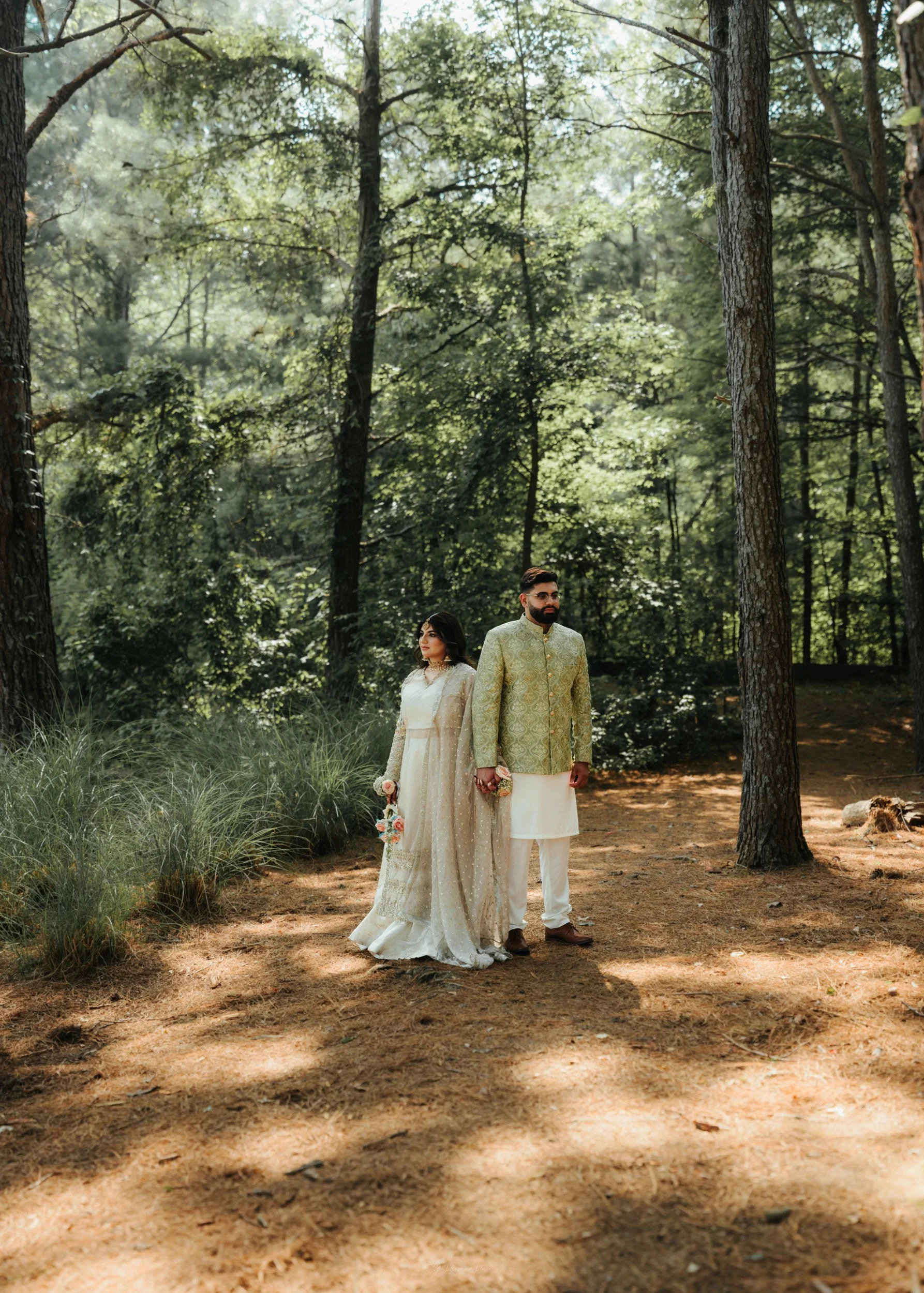 A couple in traditional Indian wedding attire standing hand-in-hand in a forest with tall trees and sunlight filtering through the leaves.