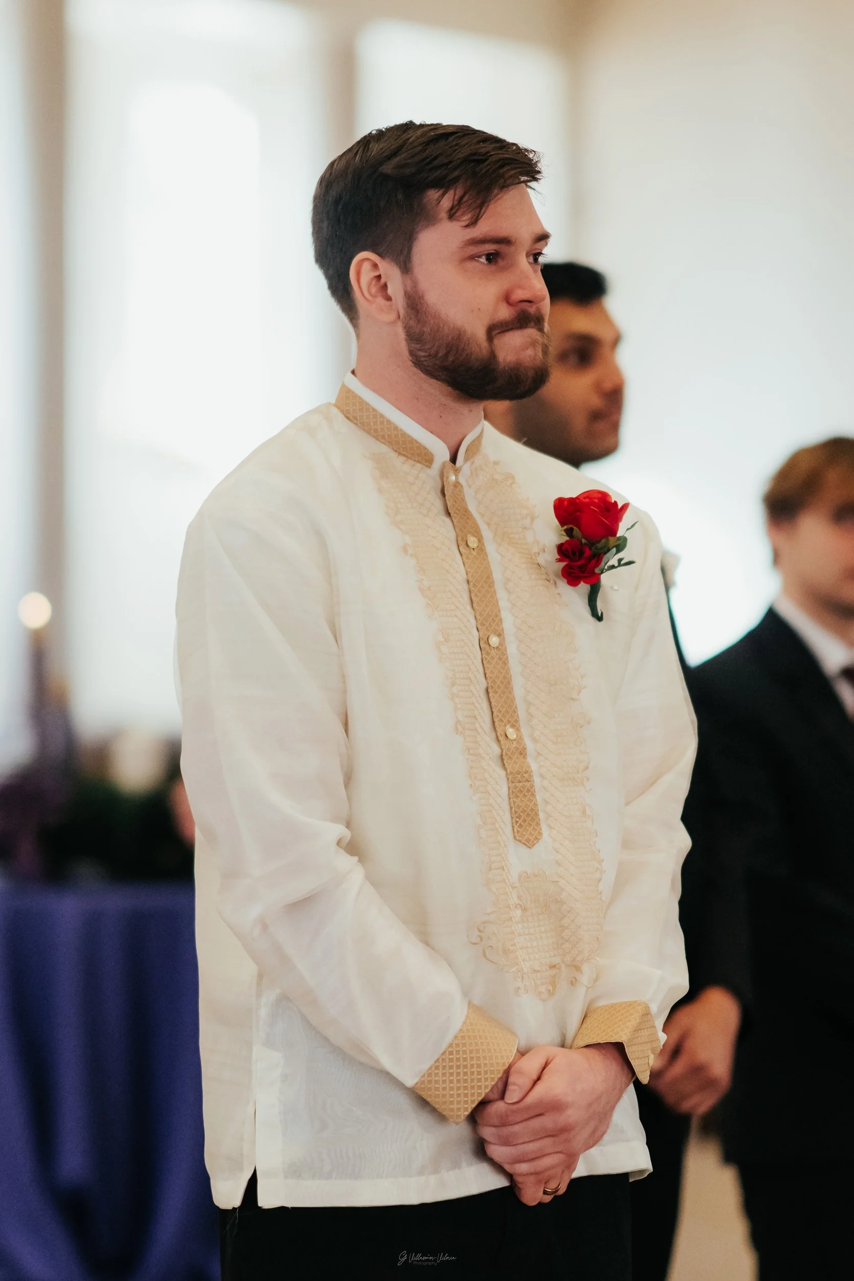 A man dressed in a traditional cream-colored Filipino Barong with gold accents, standing with his hands clasped, during a formal ceremony or wedding, with a red boutonniere on his left chest.