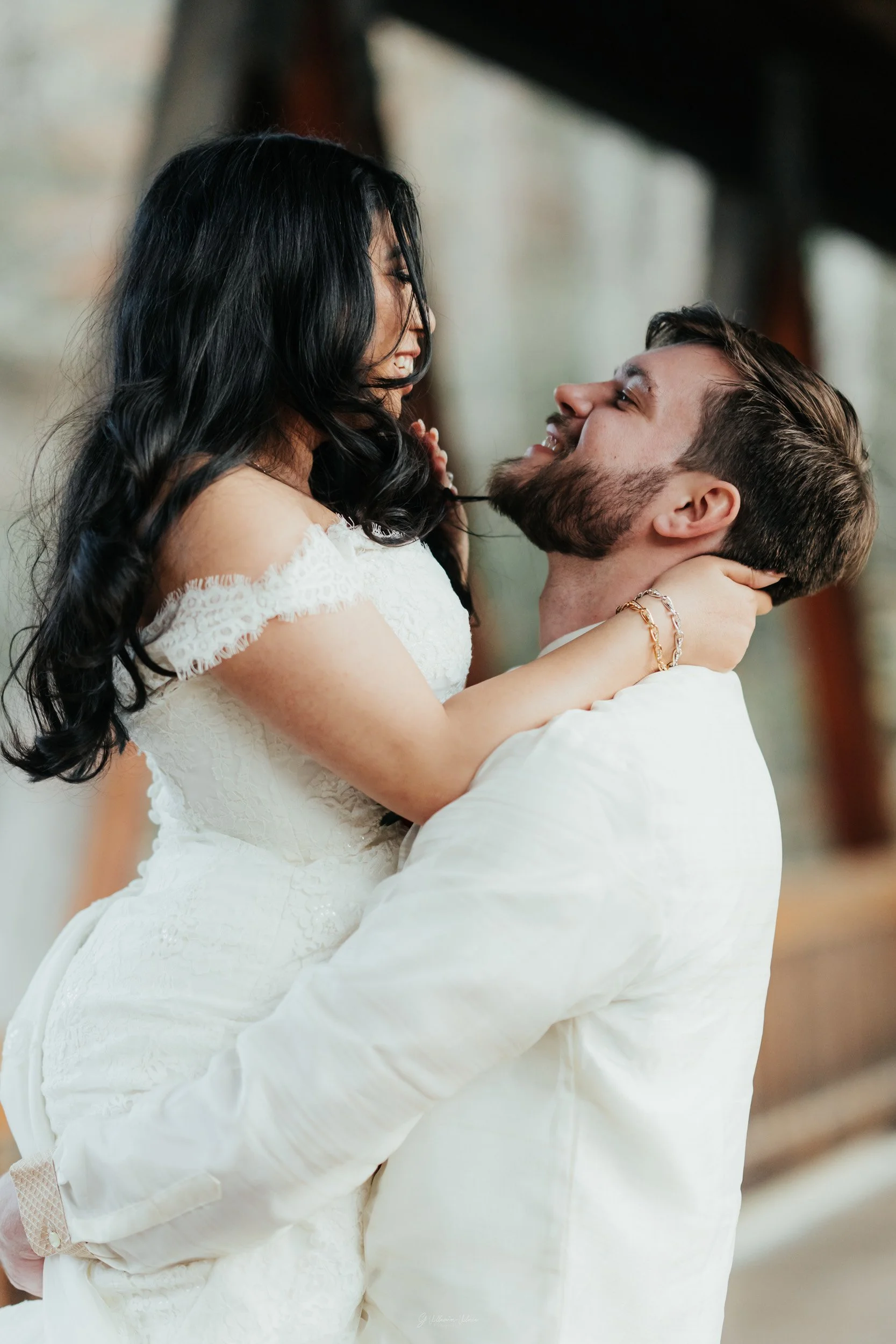 A couple, dressed in wedding attire, are smiling and looking into each other's eyes. The woman is being lifted by the man, holding her with both hands, and they appear very happy together.