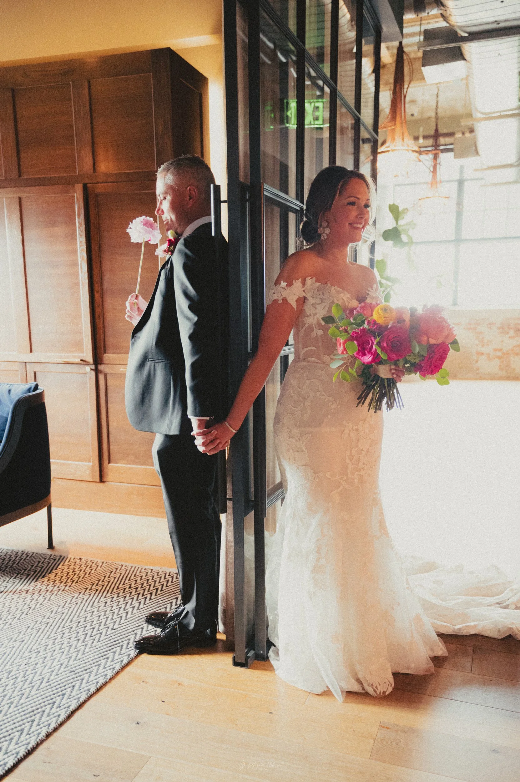 A bride and groom are standing back to back, holding hands, with their backs touching a glass door. The bride is smiling and holding a large bouquet of colorful flowers, wearing a lace wedding dress. The groom is dressed in a black suit and is holdin