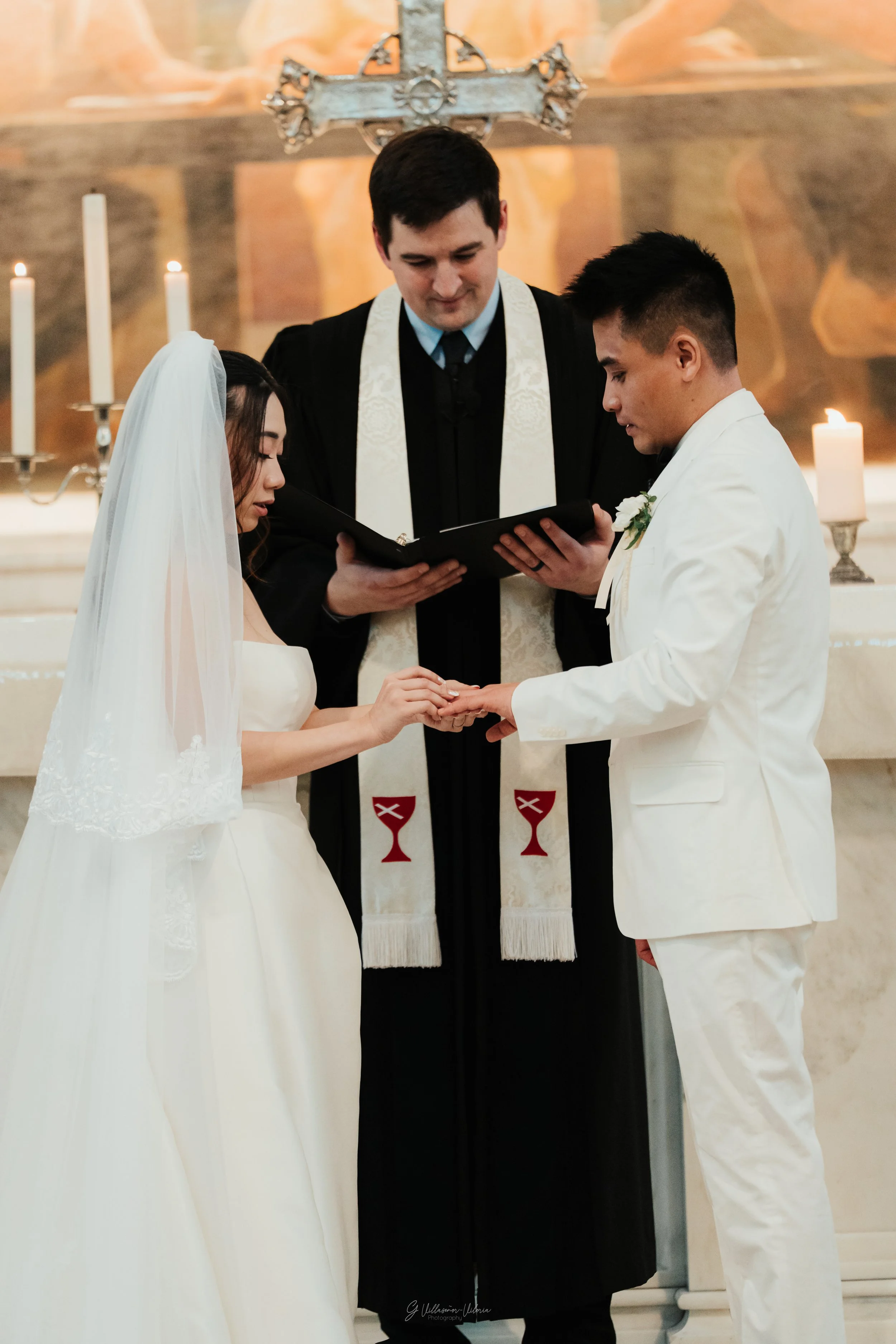 A bride and groom exchange rings during a wedding ceremony in a church, with a priest officiating and candles burning in the background.