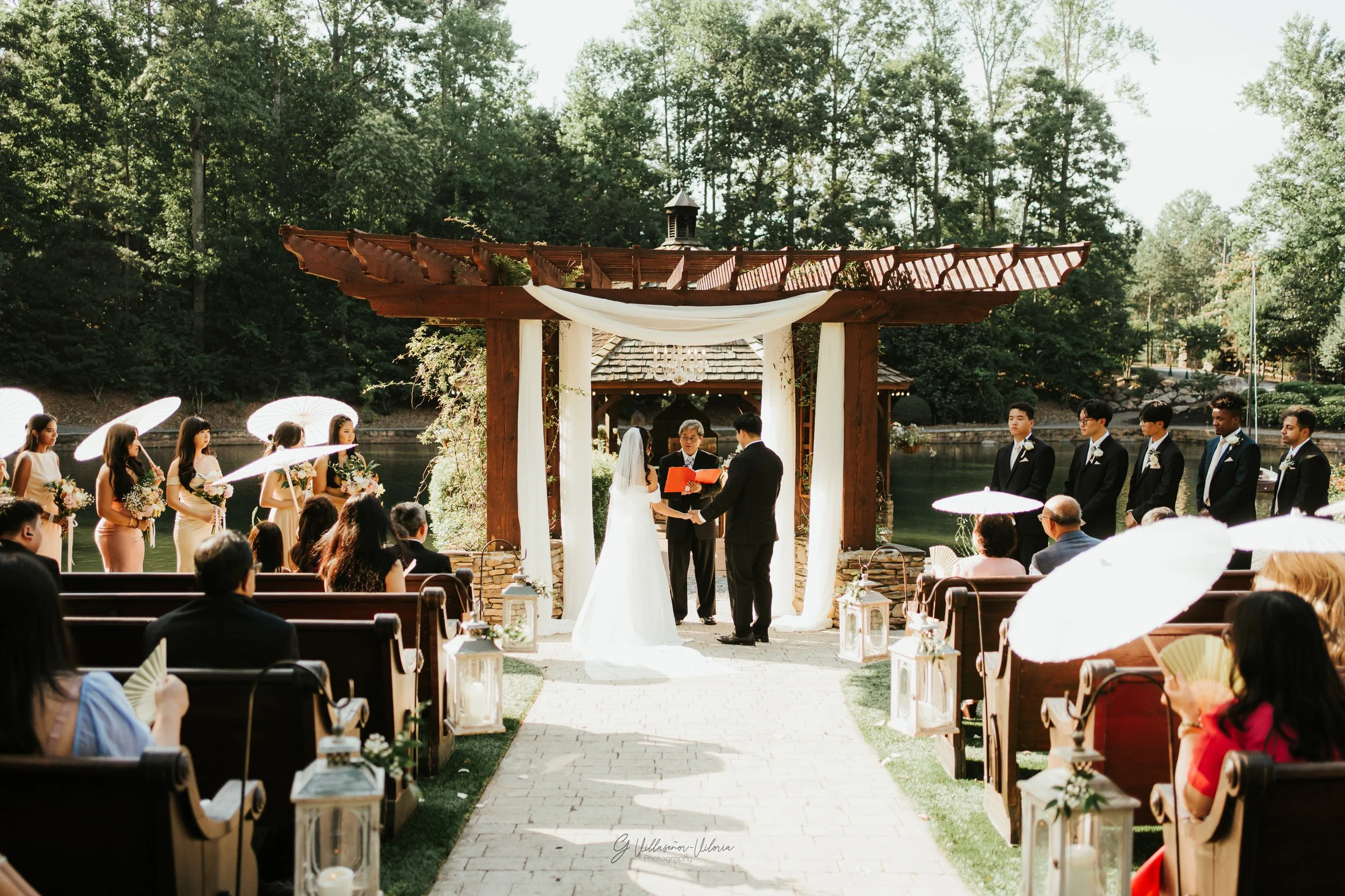 Outdoor wedding ceremony with a bride and groom exchanging vows under a wooden arbor, surrounded by bridesmaids holding umbrellas on the left and groomsmen on the right, with guests seated on benches and lanterns lining the aisle.
