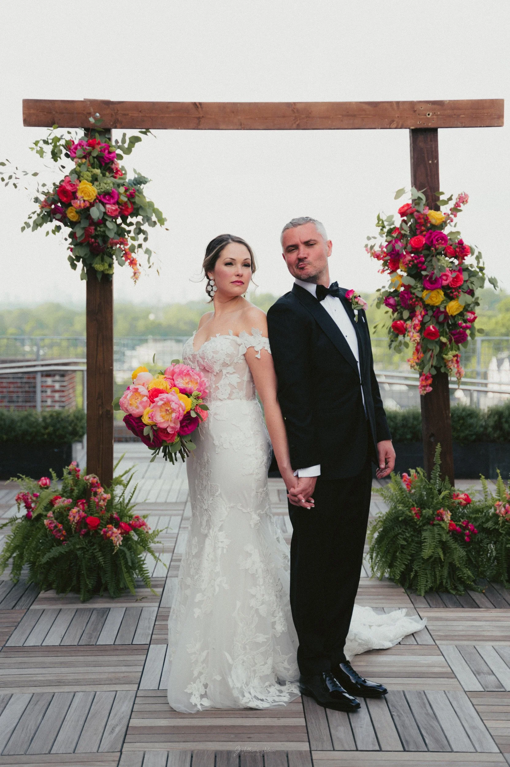 A bride and groom holding hands at a wedding ceremony outdoor, standing in front of a wooden floral arch with pink, yellow, and red flowers, on a rooftop with a city skyline background.