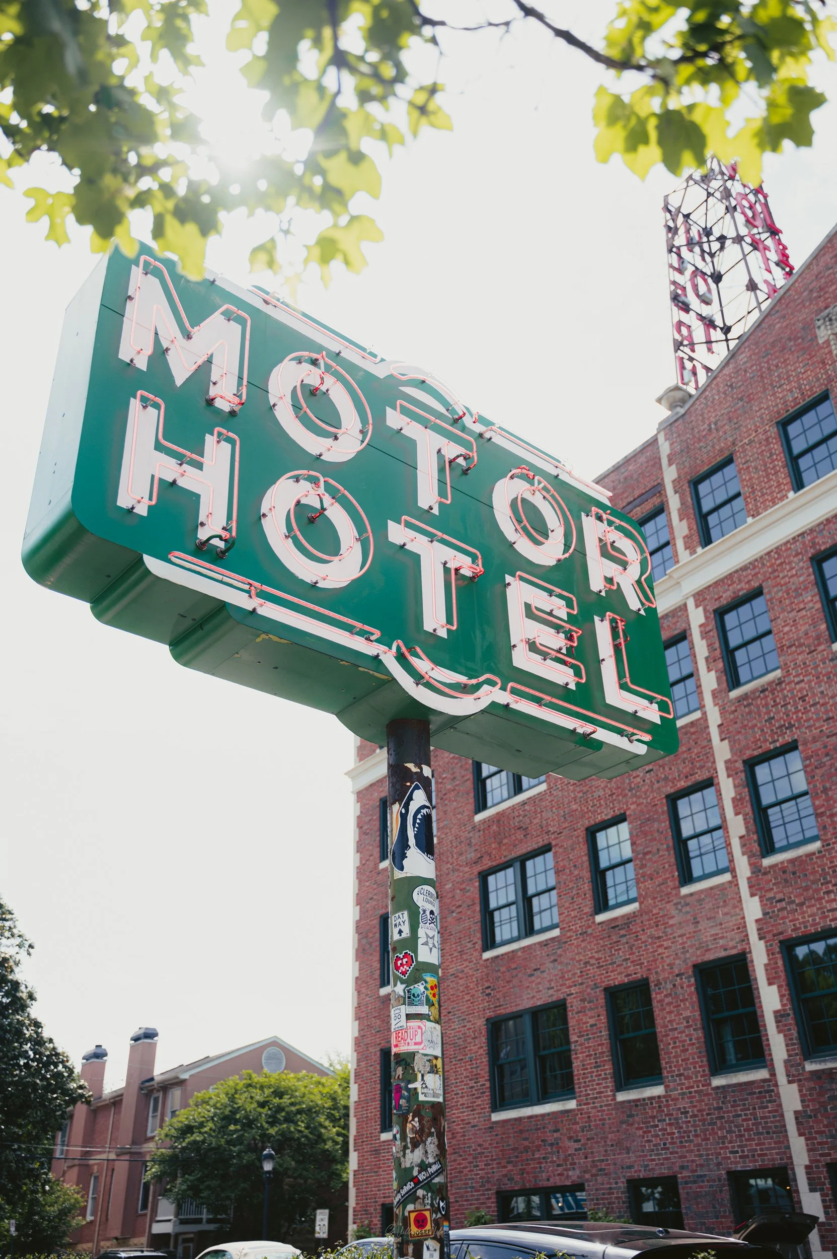 Neon sign reading 'Motor Hotel' mounted on a pole covered with stickers, with red brick building and trees in the background.