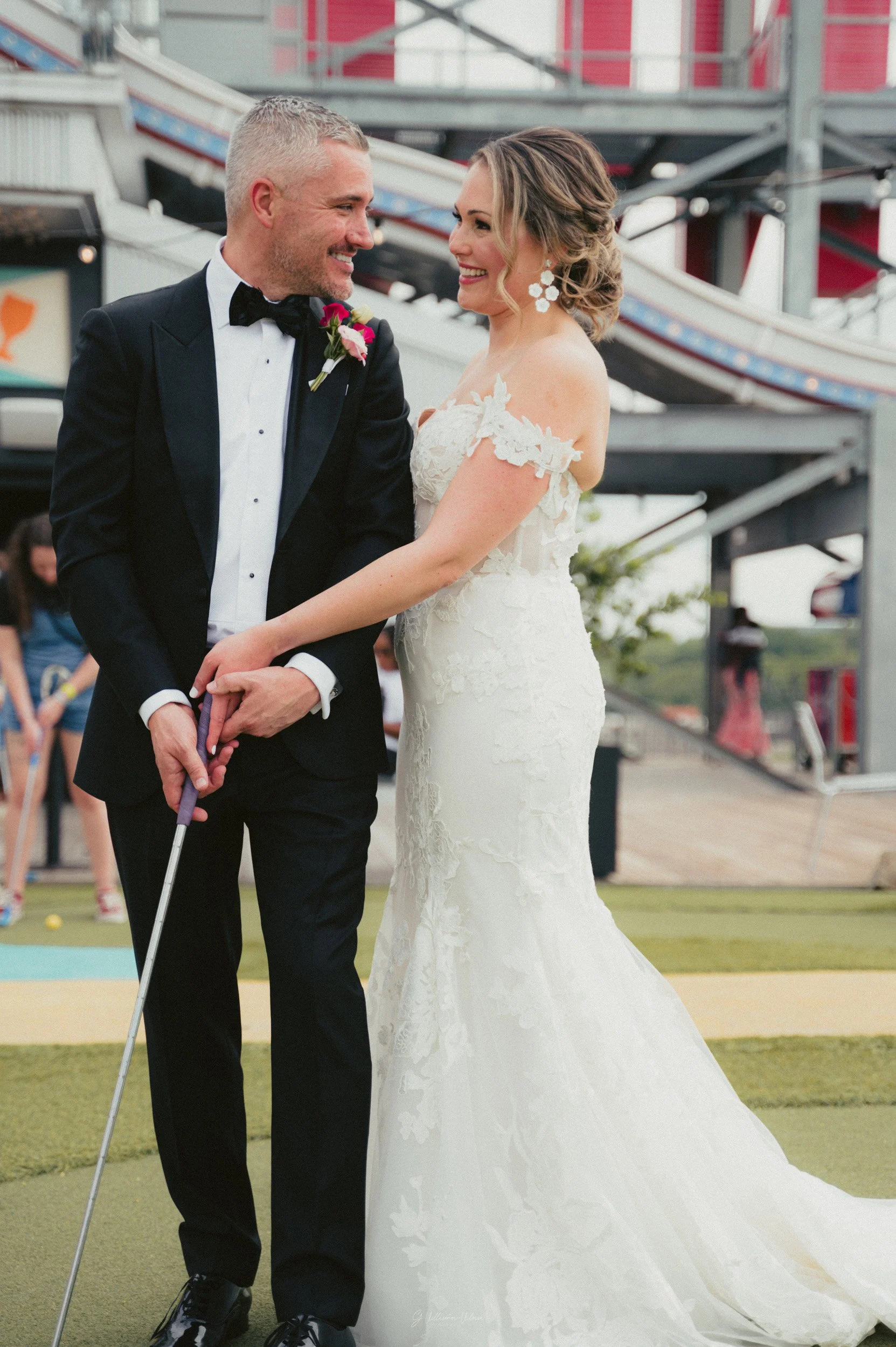 A bride and groom smiling and holding hands at a wedding, with an amusement park ride and other people in the background.
