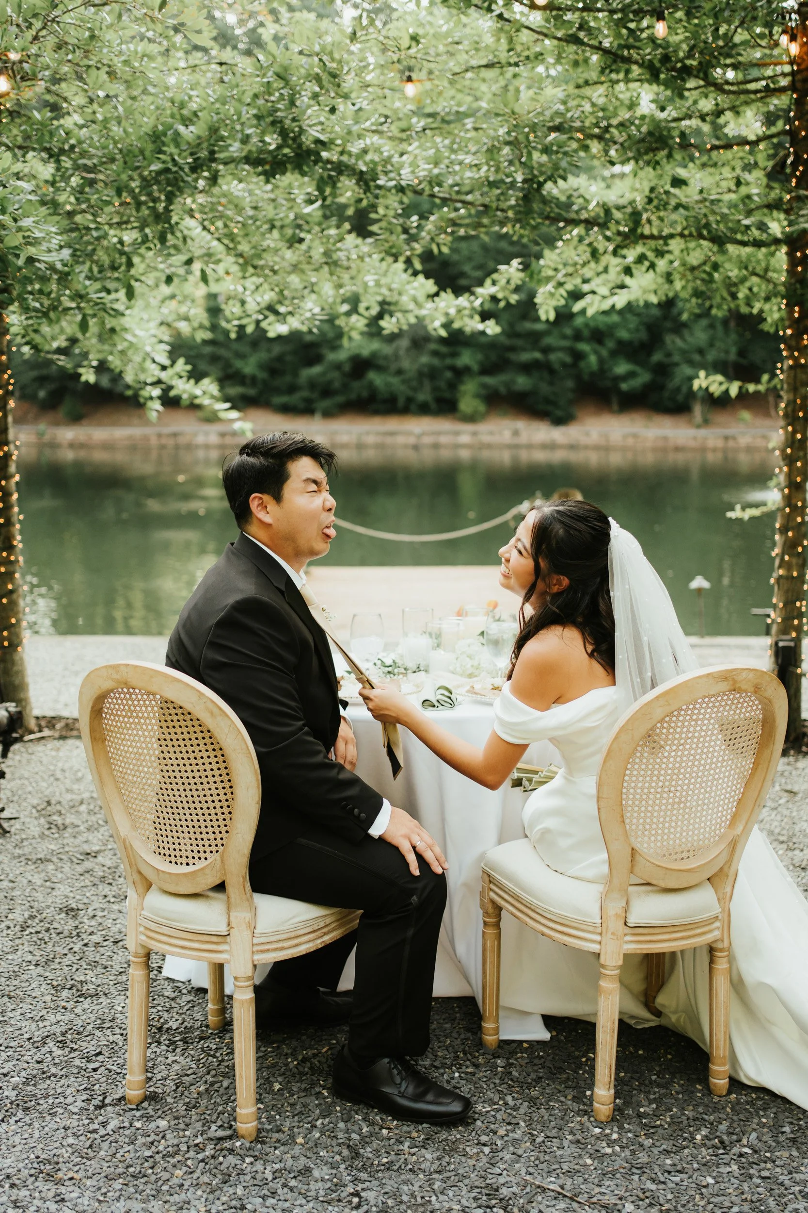 A bride and groom having fun during their wedding reception outdoors by a lake, with a tree decorated with string lights in the background.