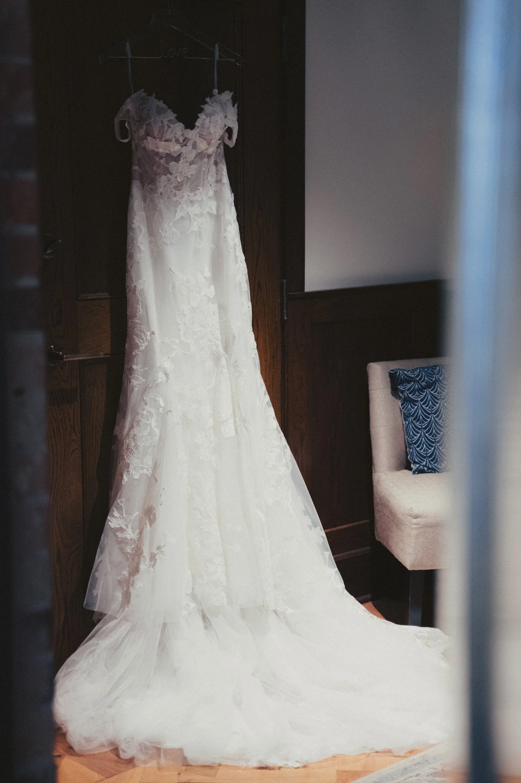 A white lace wedding dress hanging on a dark wooden wall with a 'Love' hanger, next to a beige chair with a blue patterned pillow.