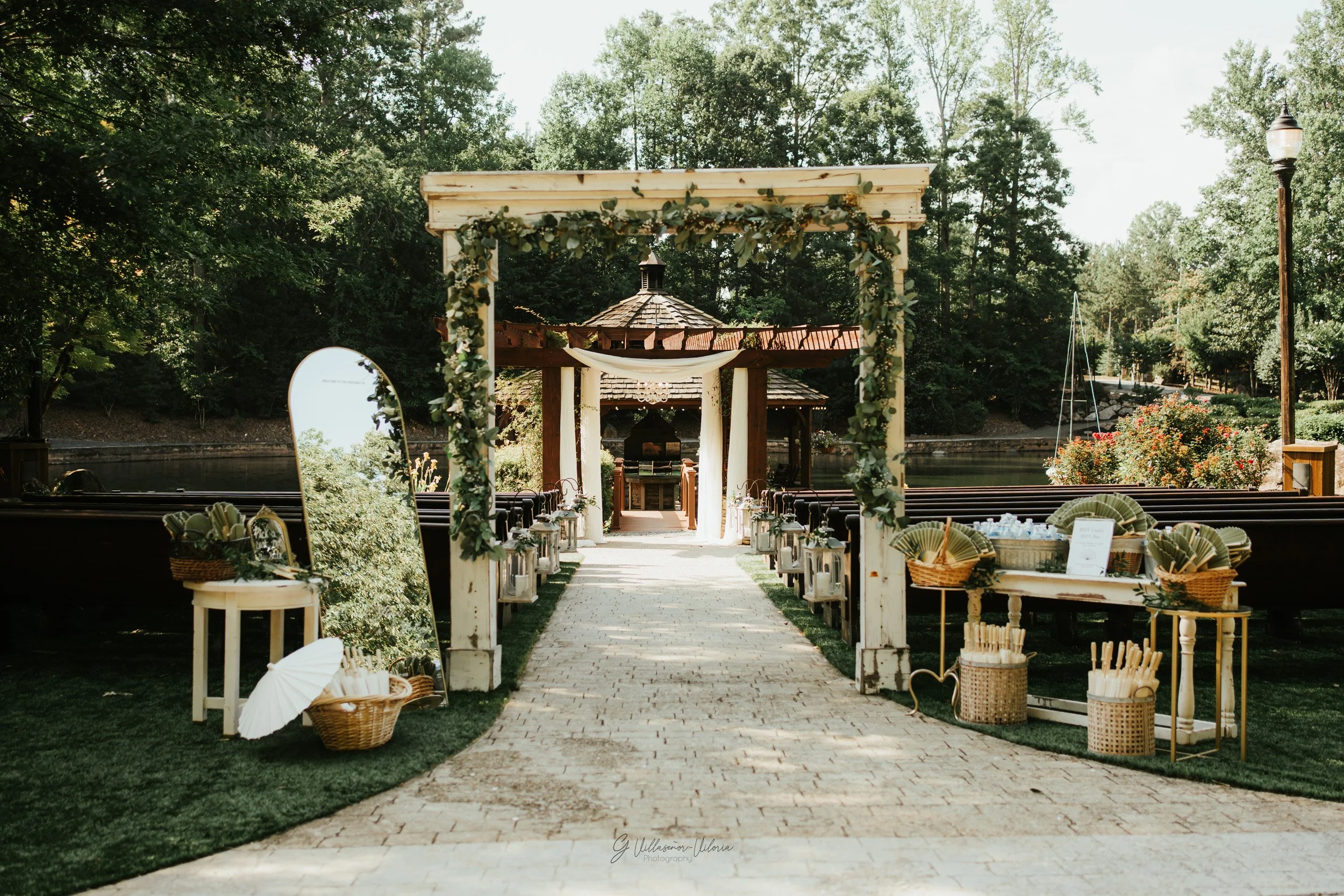 Outdoor wedding setup with a pathway, wooden arch decorated with greenery, and seating area surrounded by trees and lake.