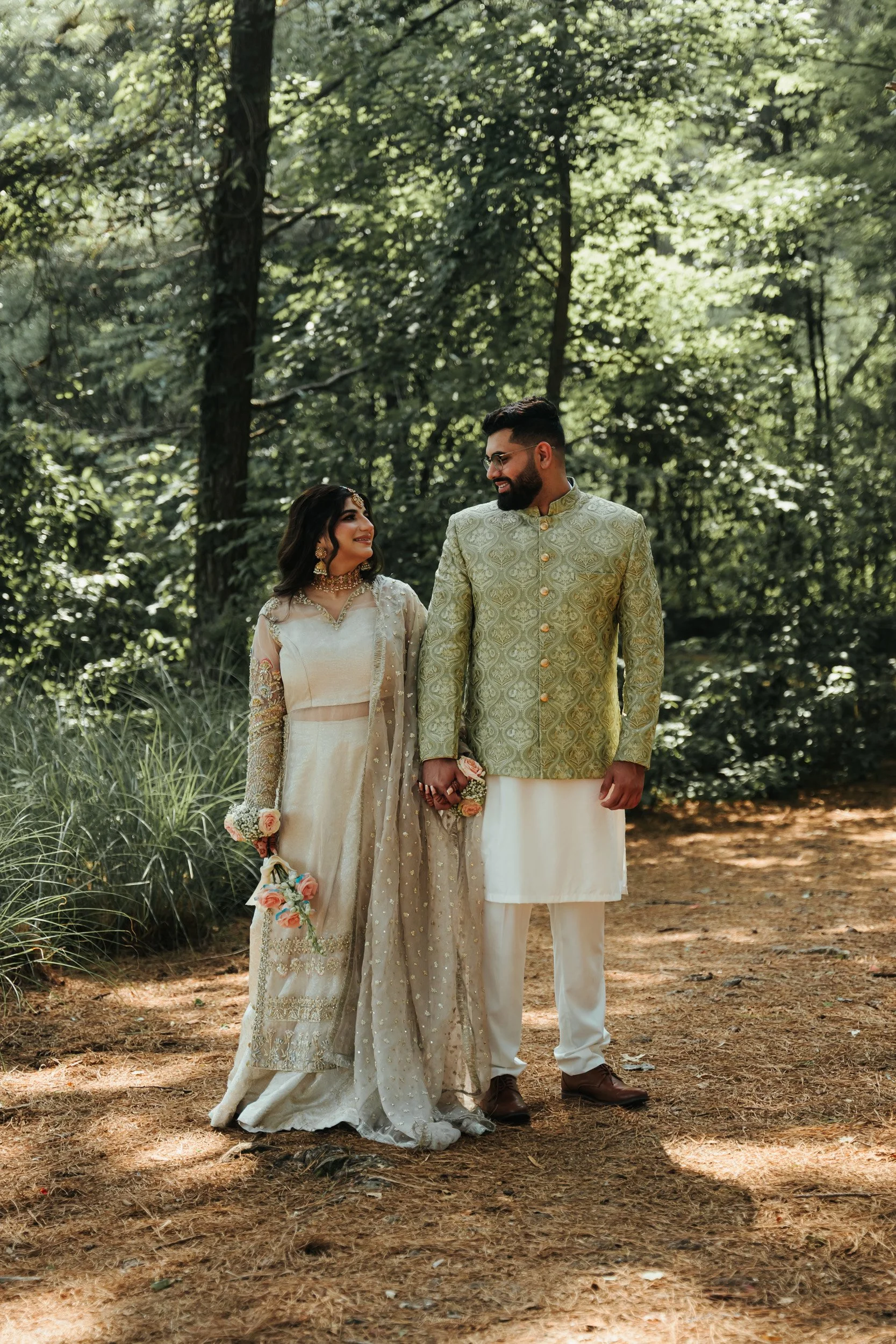 A couple dressed in traditional South Asian wedding attire standing outdoors in a forested area, holding hands and looking at each other.