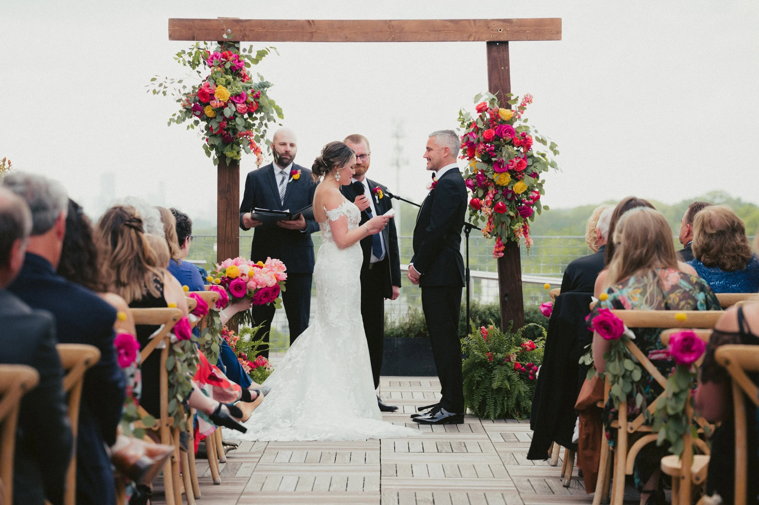 A wedding ceremony taking place outdoors under a wooden arch decorated with vibrant pink, red, and yellow flowers. The bride and groom are facing each other, with the bride reading vows and the groom listening. The bride wears a white lace gown, and 