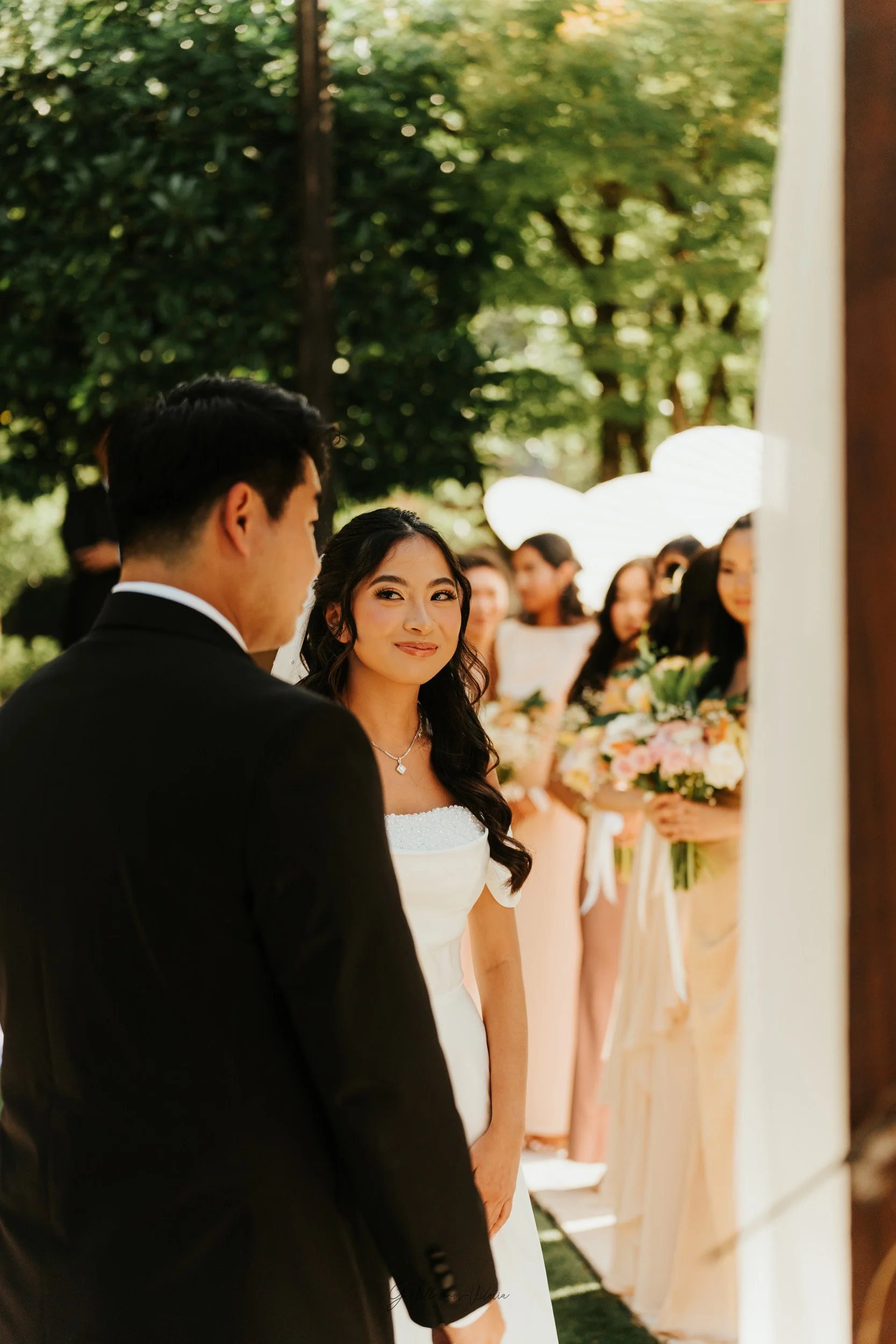 A bride with dark wavy hair and a silver necklace looks at her groom during their outdoor wedding ceremony, with bridesmaids holding bouquets in the background.