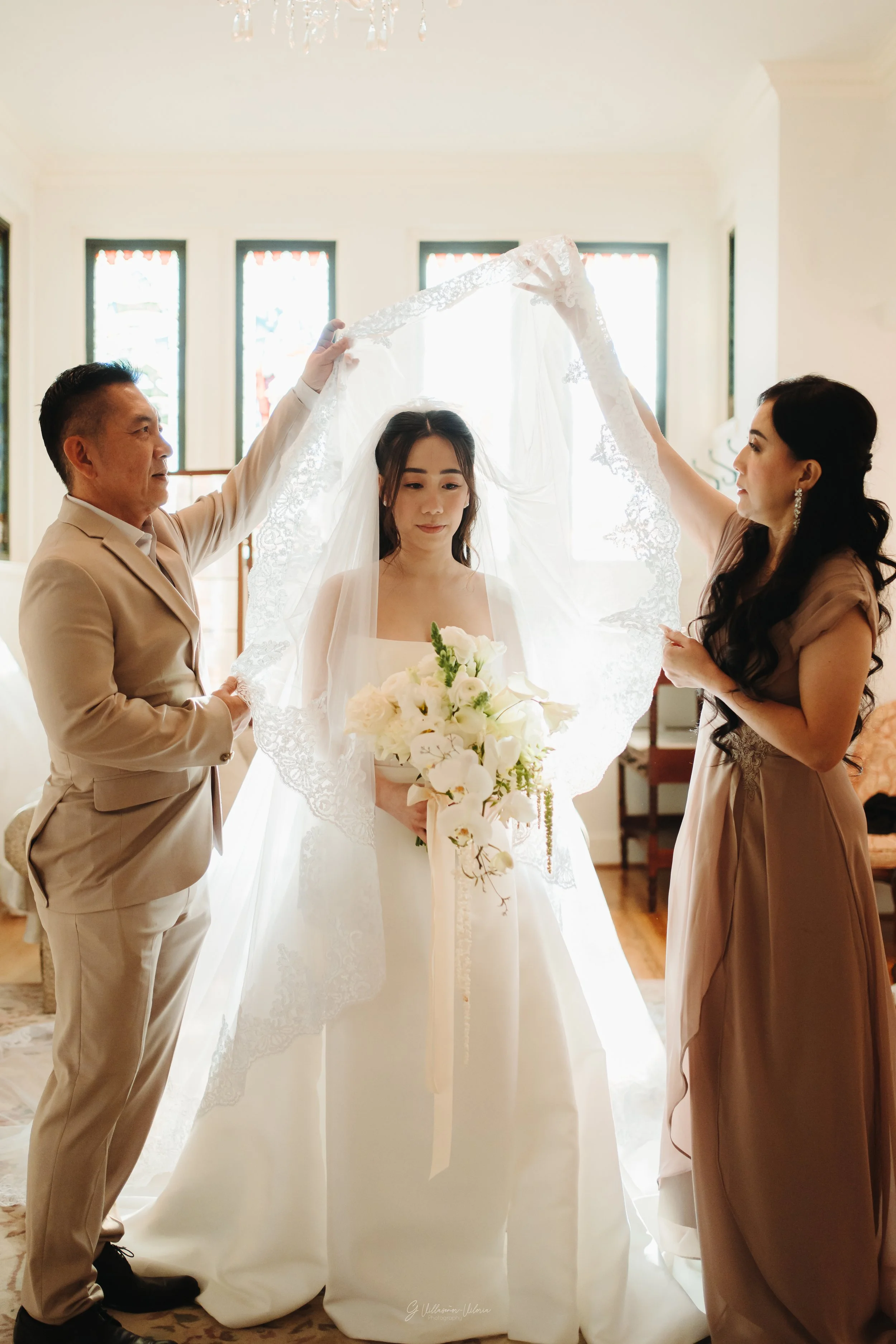 A bride in a white wedding gown is standing with a bouquet of white flowers, while two people, possibly her parents, hold a lace veil over her head indoors, with sunlight streaming through windows in the background.