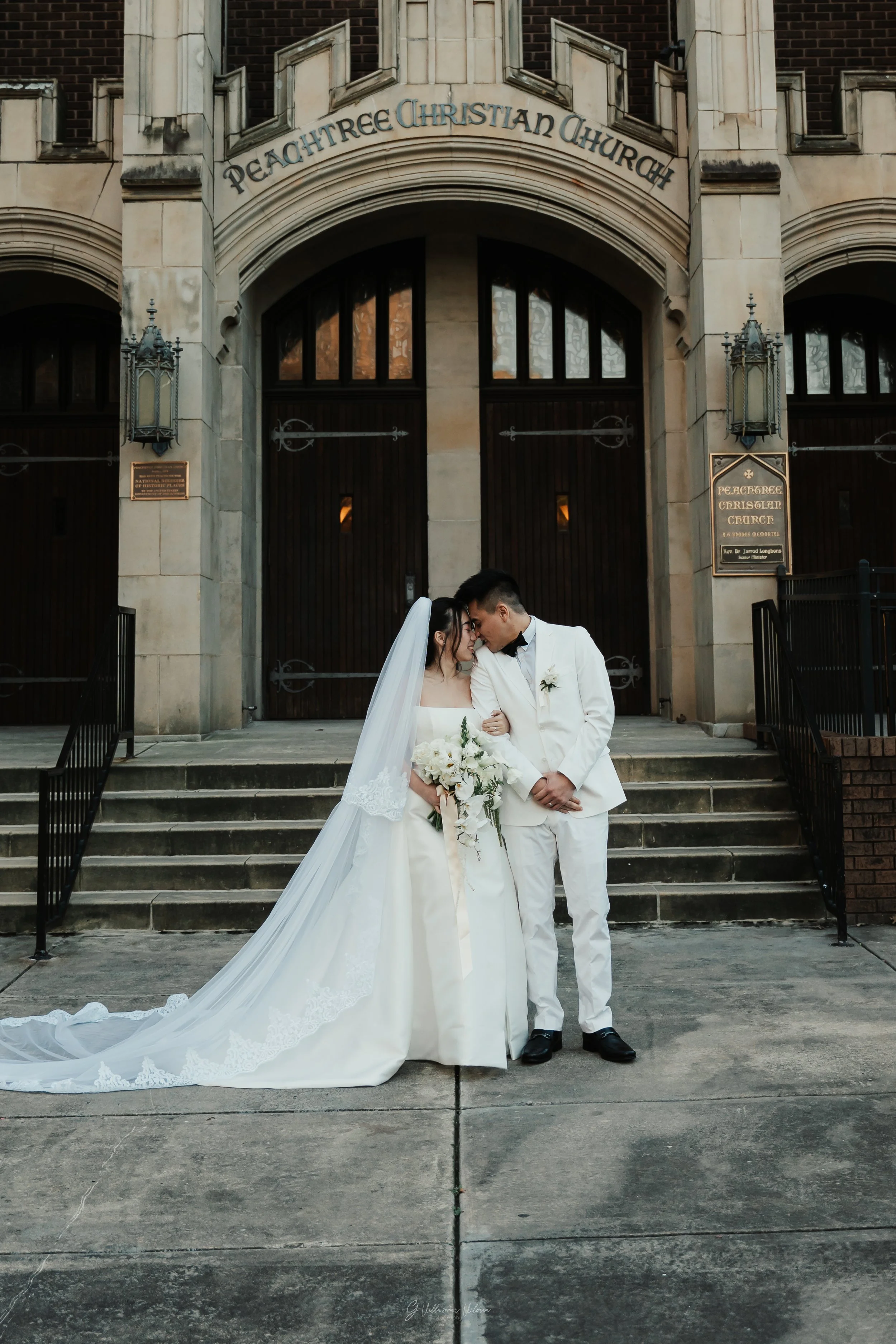 A bride and groom in wedding attire posing for a photo on the steps outside Peachtree Christian Church.