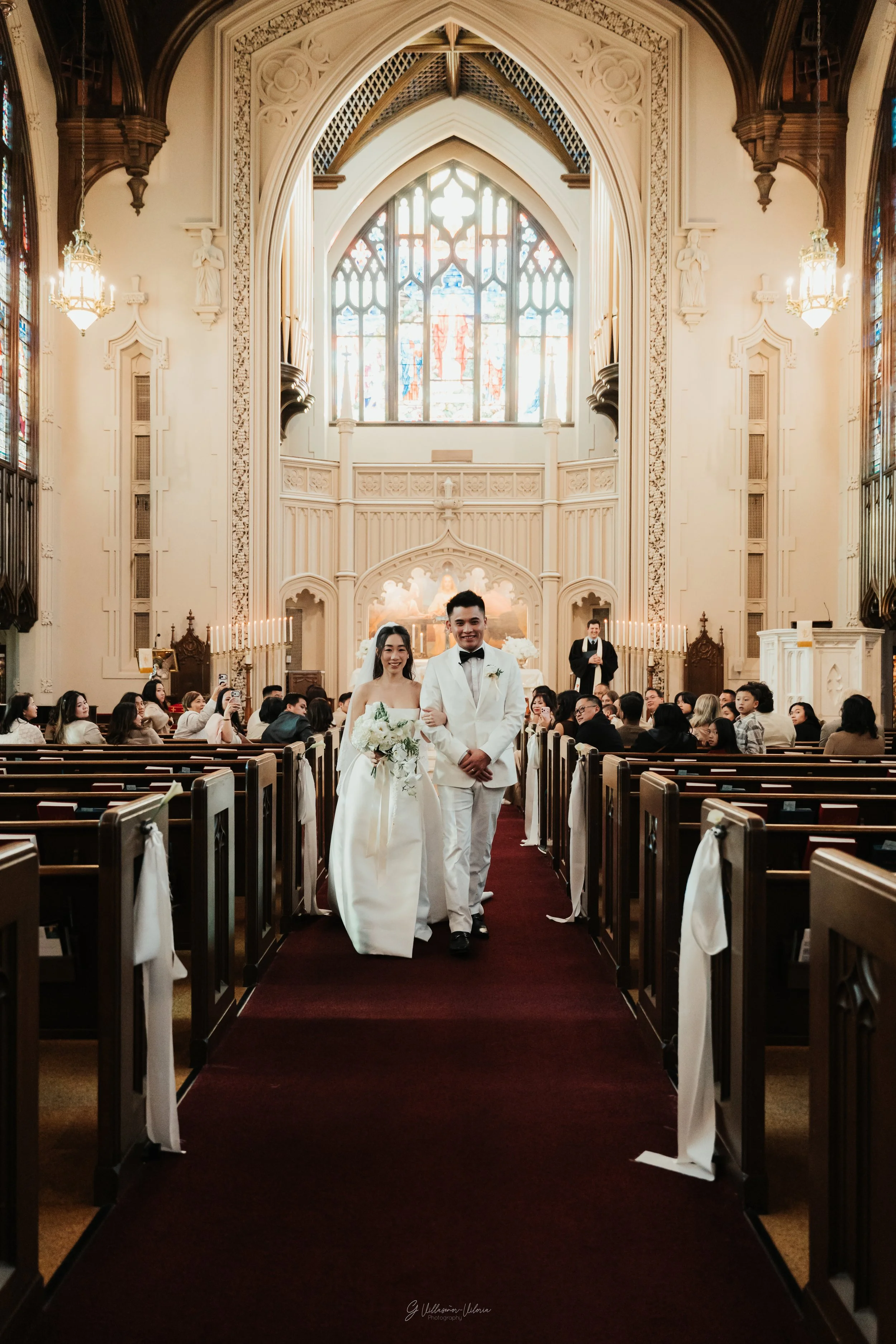 A bride and groom walk down the aisle inside a church with stained glass windows after their wedding ceremony. The bride is holding a bouquet of white flowers, and the groom is dressed in a white tuxedo with a black bow tie. Guests are seated in the 