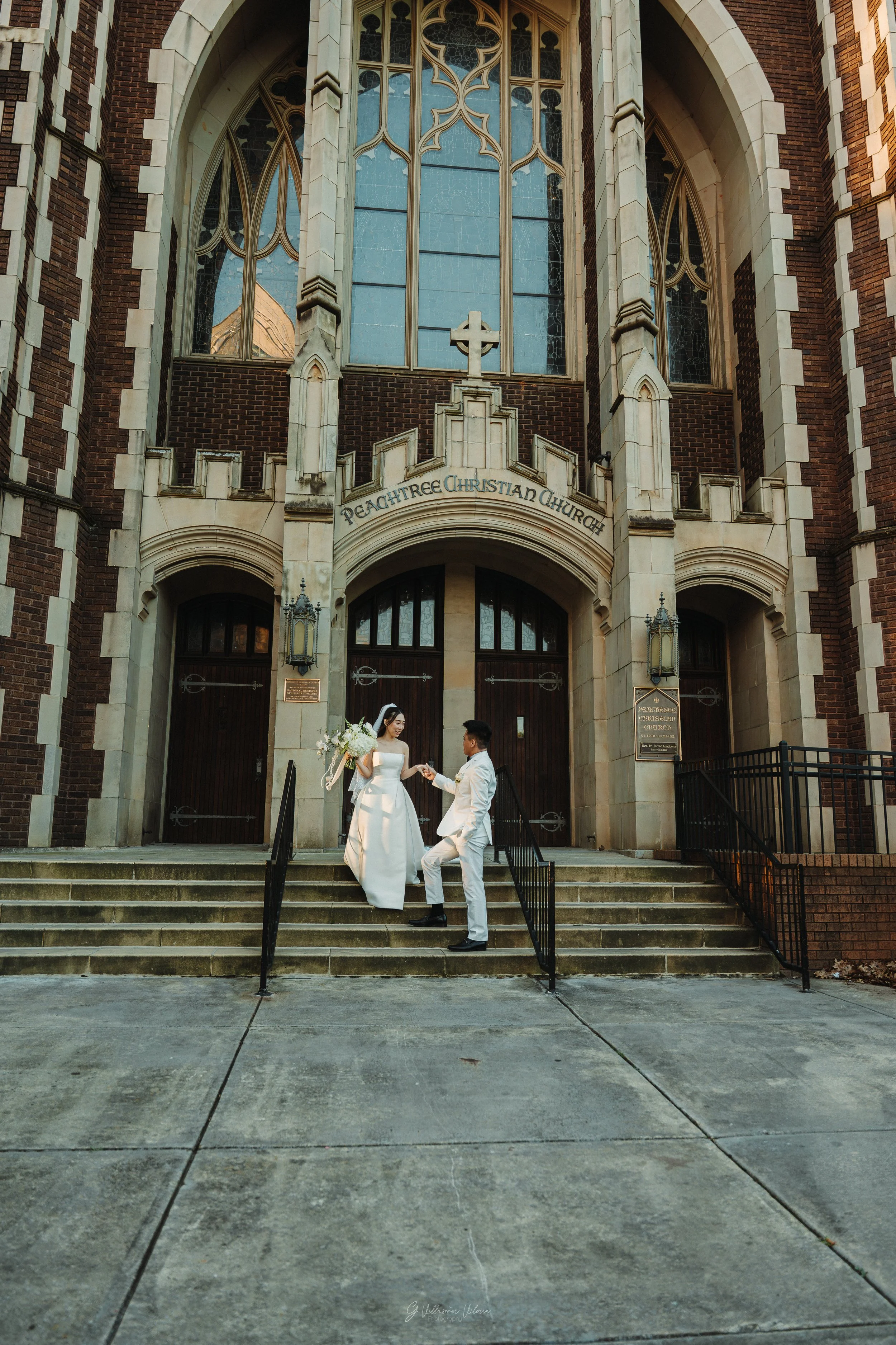 Bride and groom on the steps of Peachtree Christian Church, with the bride holding a bouquet and the groom dressed in white, exchanging rings or a handshake.
