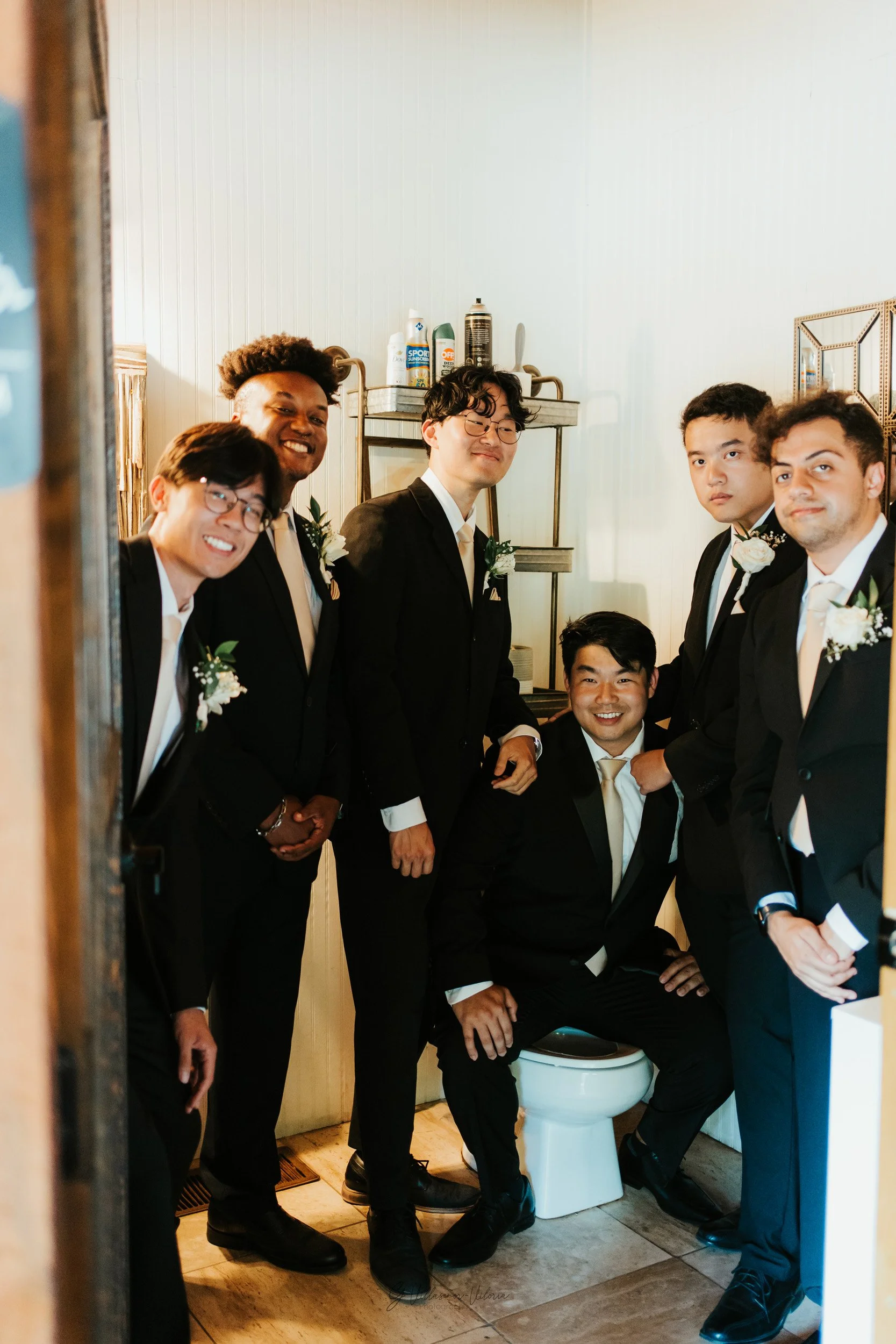 Group of seven men dressed in formal suits gathered in a small bathroom with cream-colored walls, some smiling and posing for the camera, one sitting on the toilet, in a wedding or formal event setting.