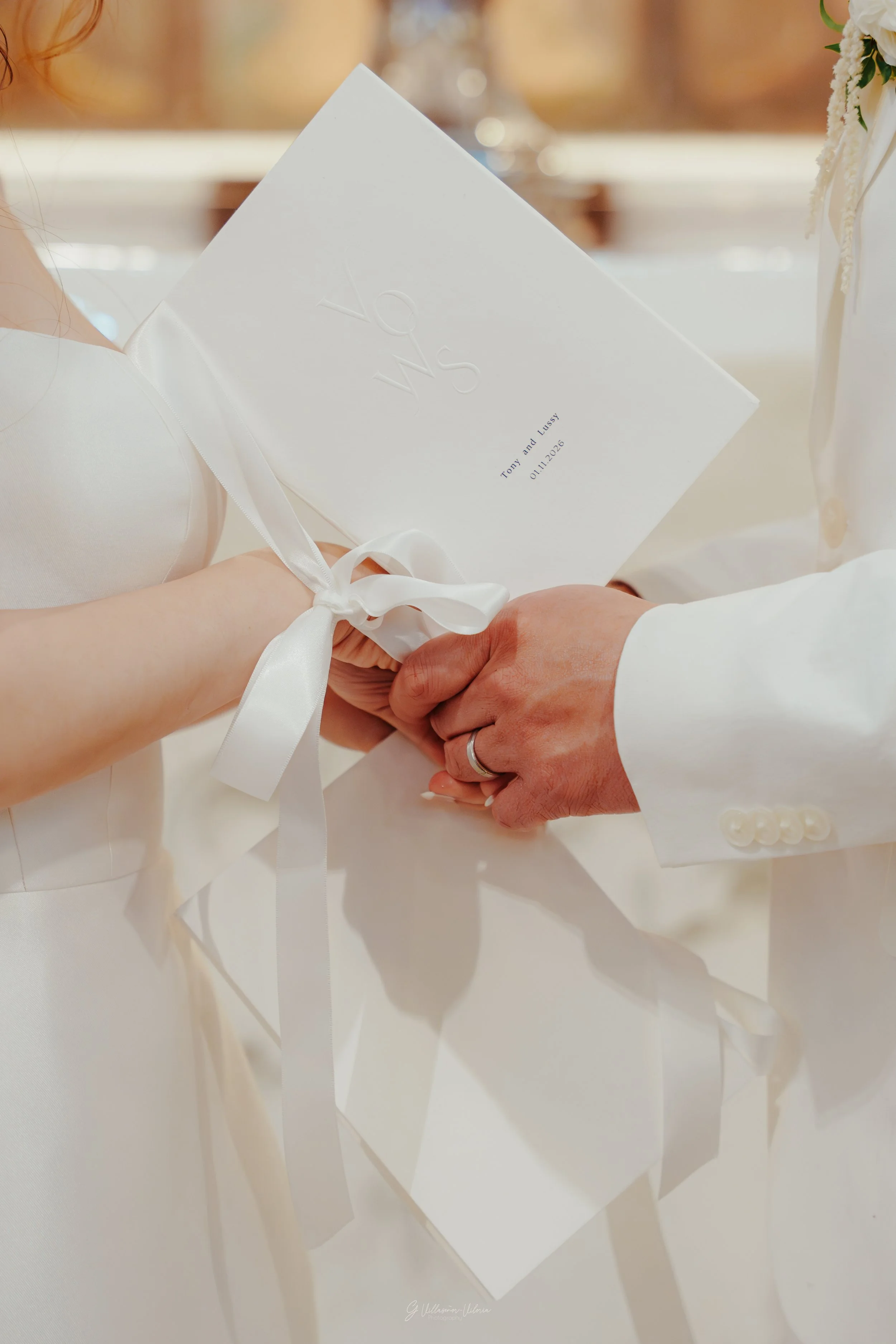 Close-up of a wedding ceremony where a bride and groom hold hands, with the bride holding a white wedding program for Tom and Lussy, dated 01.11.2026.