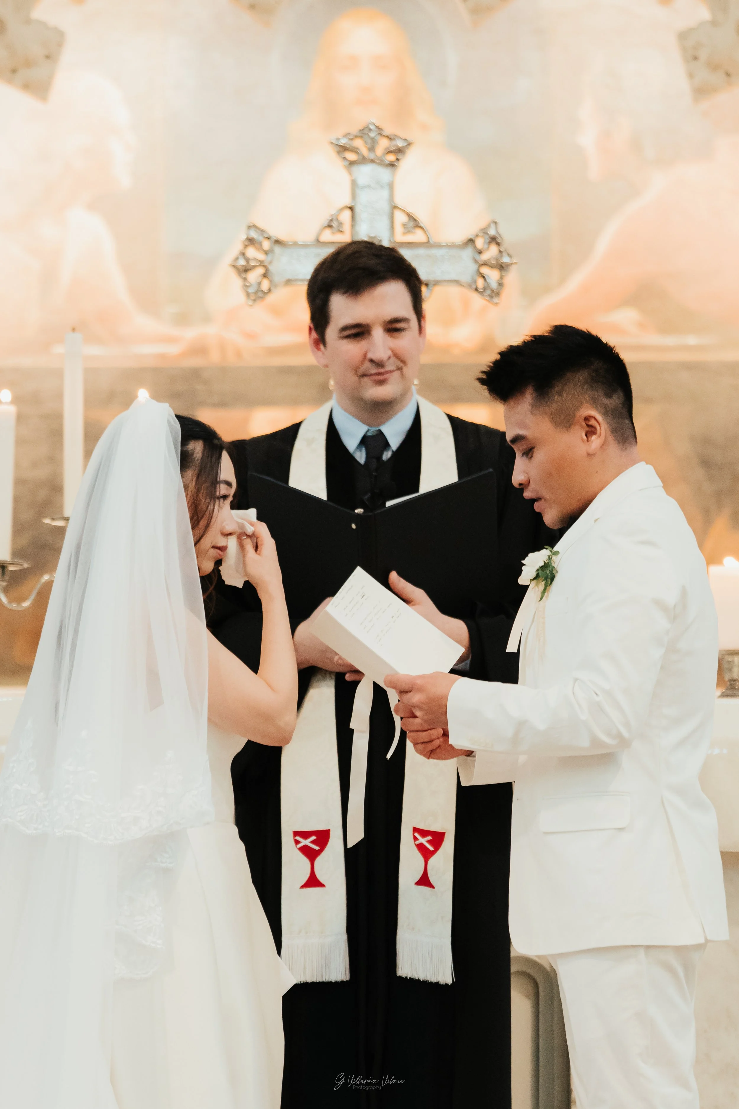 A bride and groom exchanging rings during their wedding ceremony in a church, with a priest officiating in the background, religious symbols and candles present.