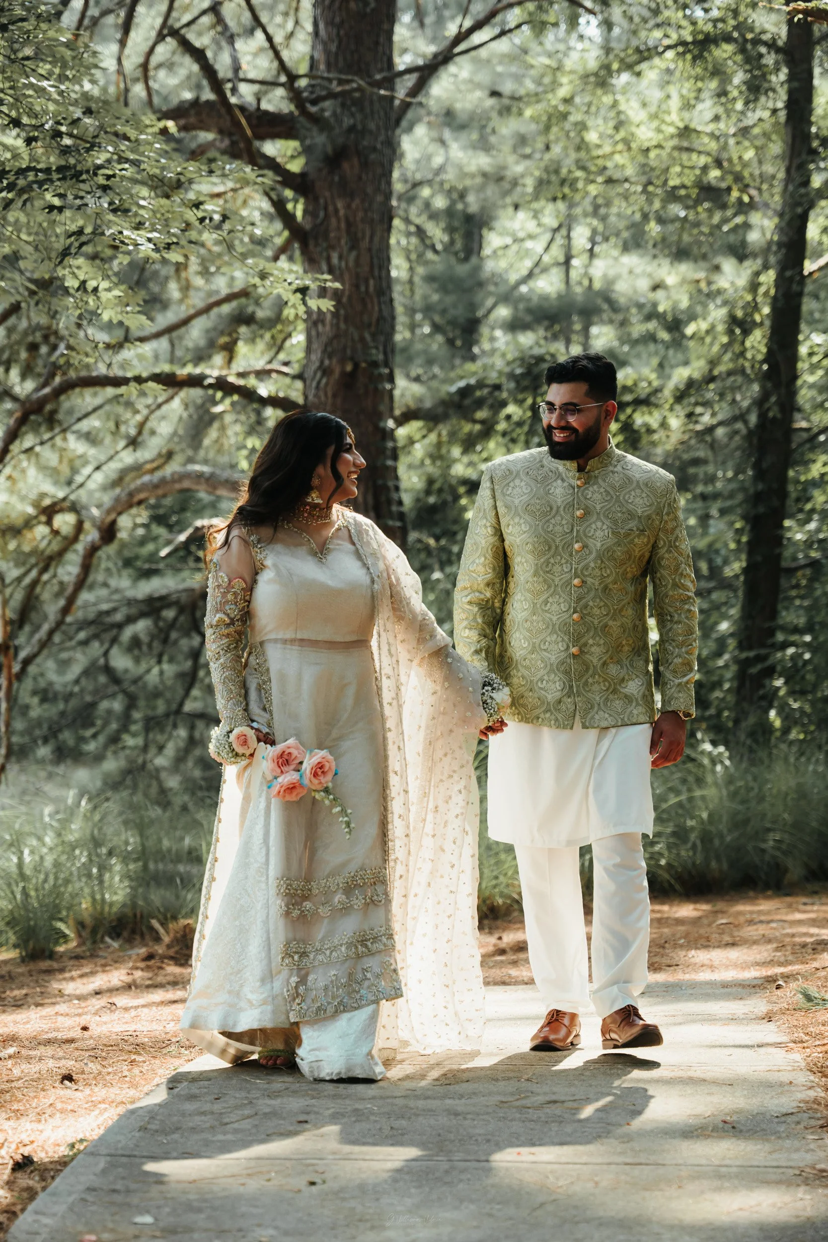 A bride and groom dressed in traditional Indian wedding attire walking together in a forest.