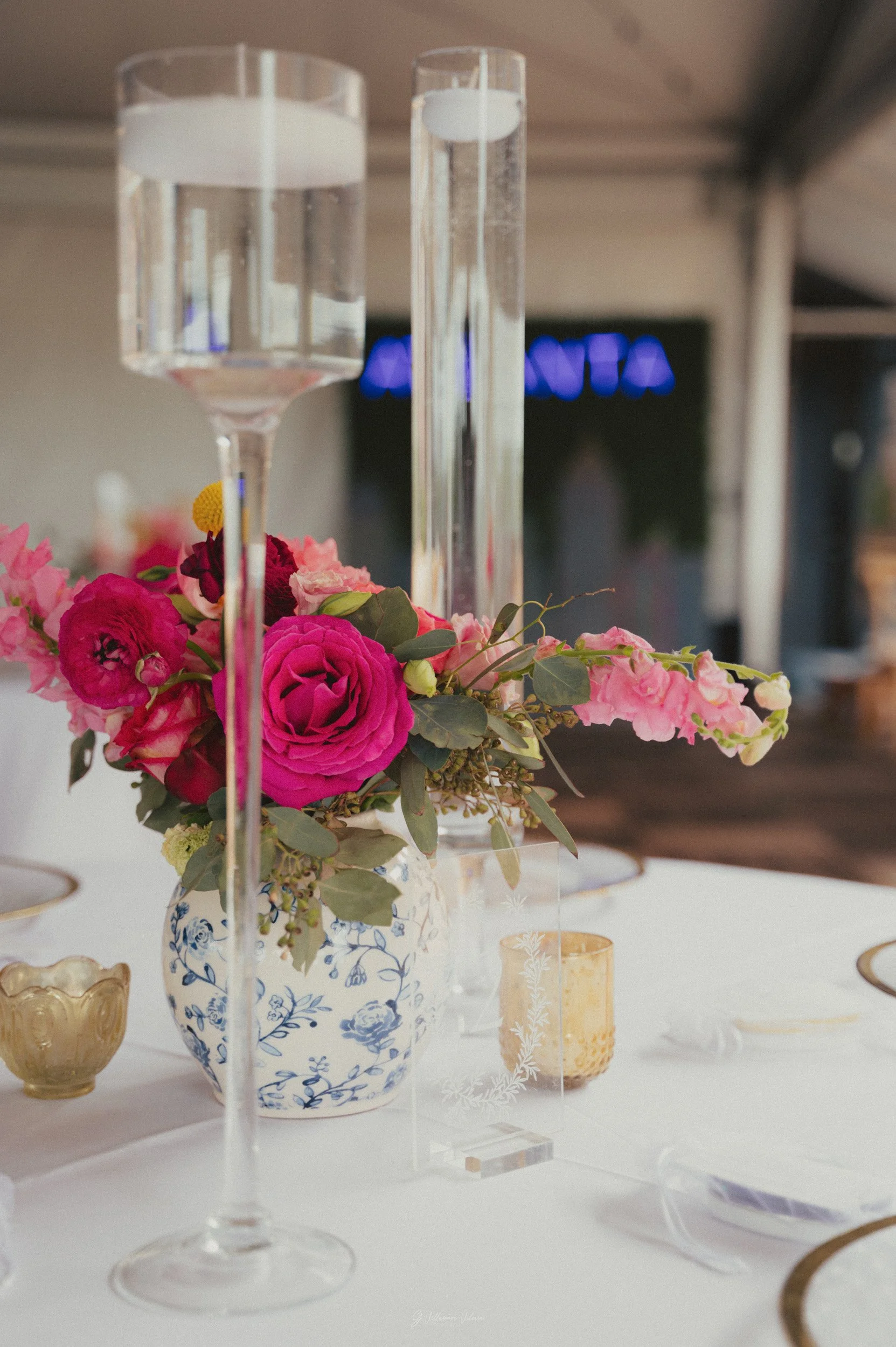 Elegant table centerpiece featuring a floral arrangement with pink and red flowers in a blue and white vase, flanked by tall glass candleholders, on a white table with gold-rimmed plates and golden candle holders.
